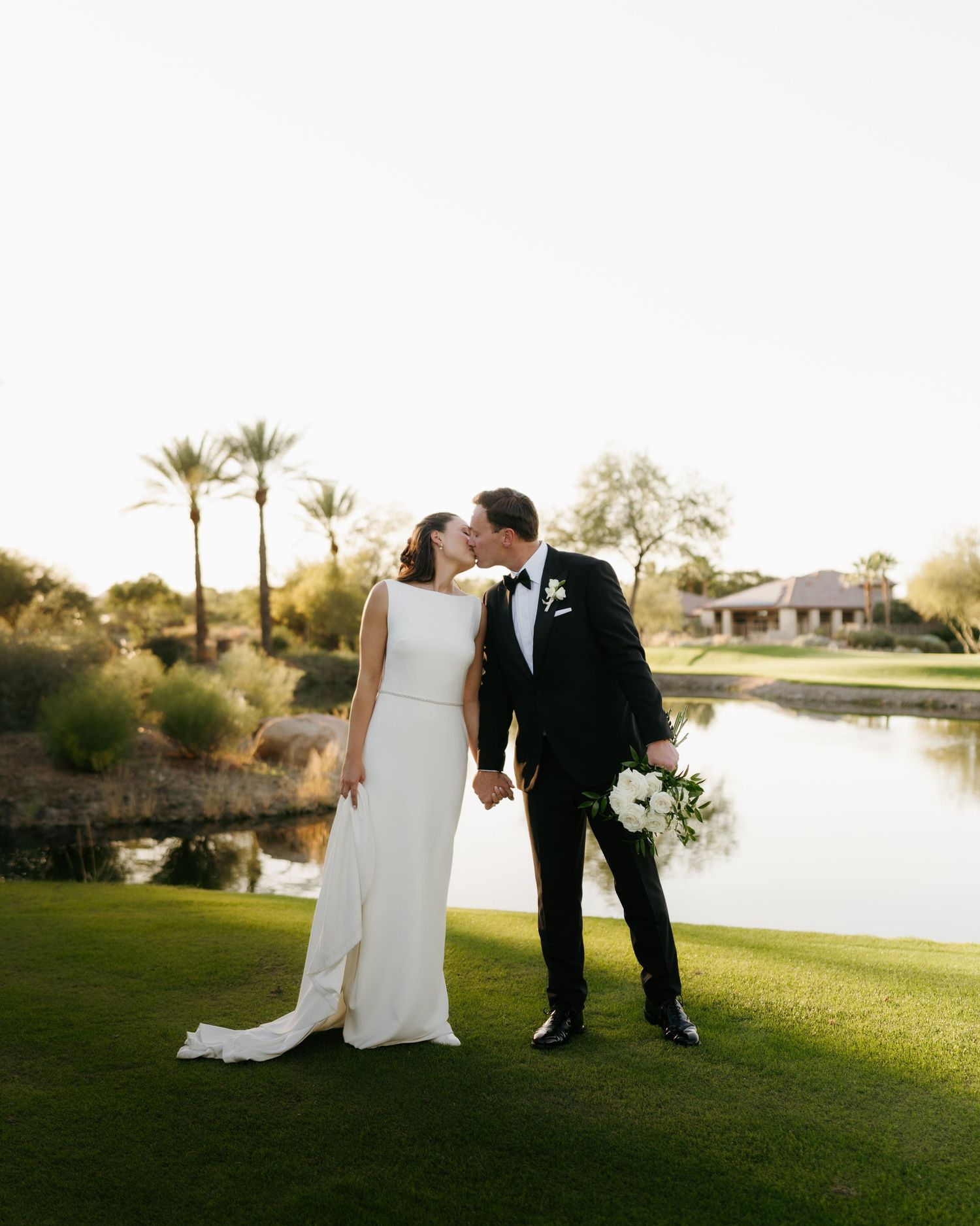 Bride and groom kissing on a golf course with palm trees and a pond at sunset.