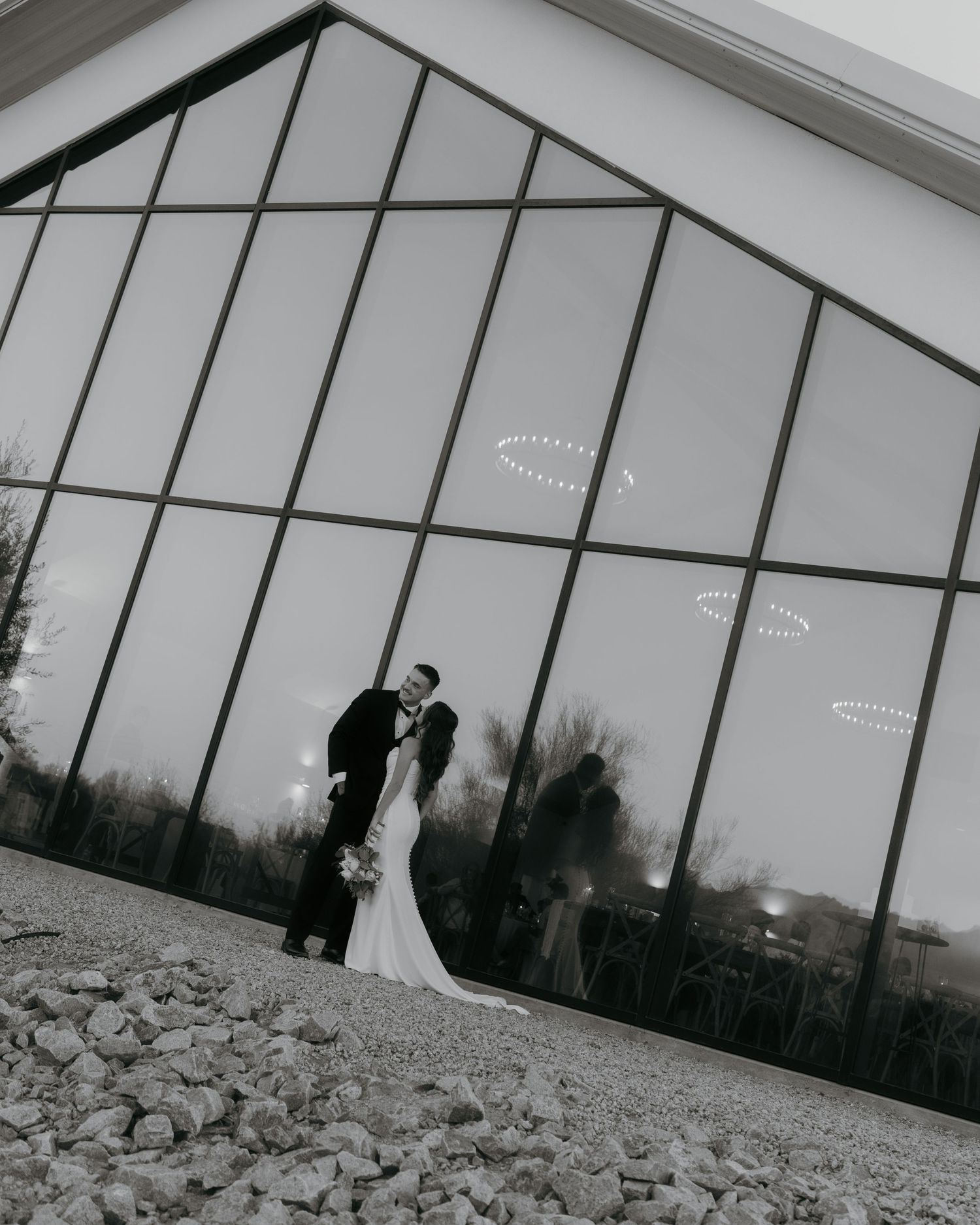 Black and white wedding photo of bride and groom kissing in front of large glass barn windows.