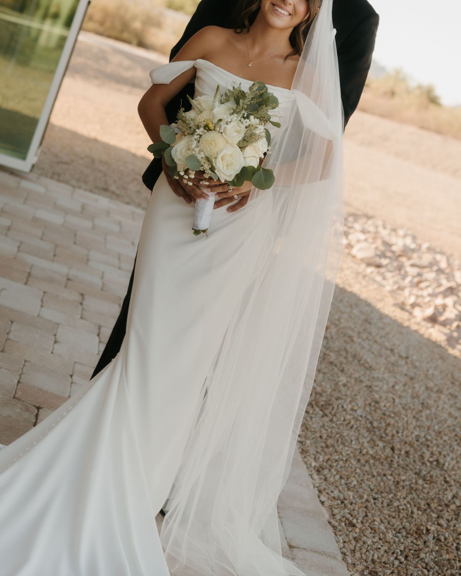 Bride in off-shoulder white gown with veil holding white rose bouquet outdoors on a sunny day.