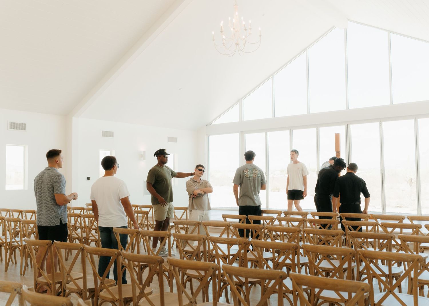 Groomsmen standing in a bright white wedding chapel with wooden chairs and a chandelier before the ceremony.