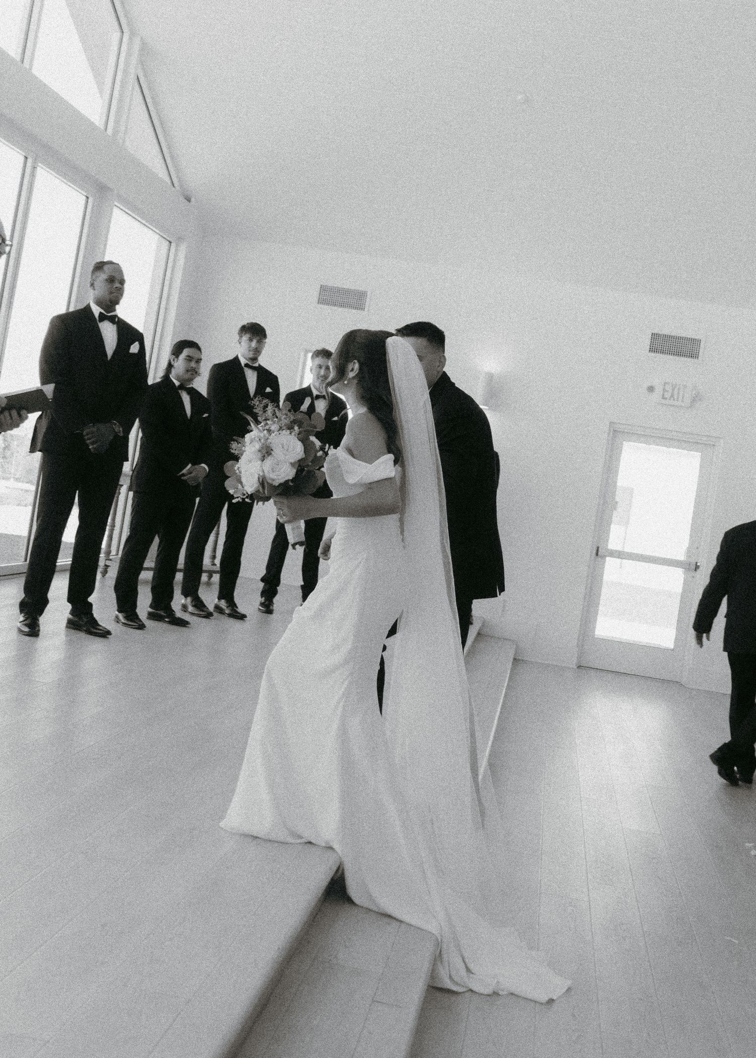Black and white photo of a bride in a white gown and veil holding a bouquet, walking toward groomsmen in a bright venue.