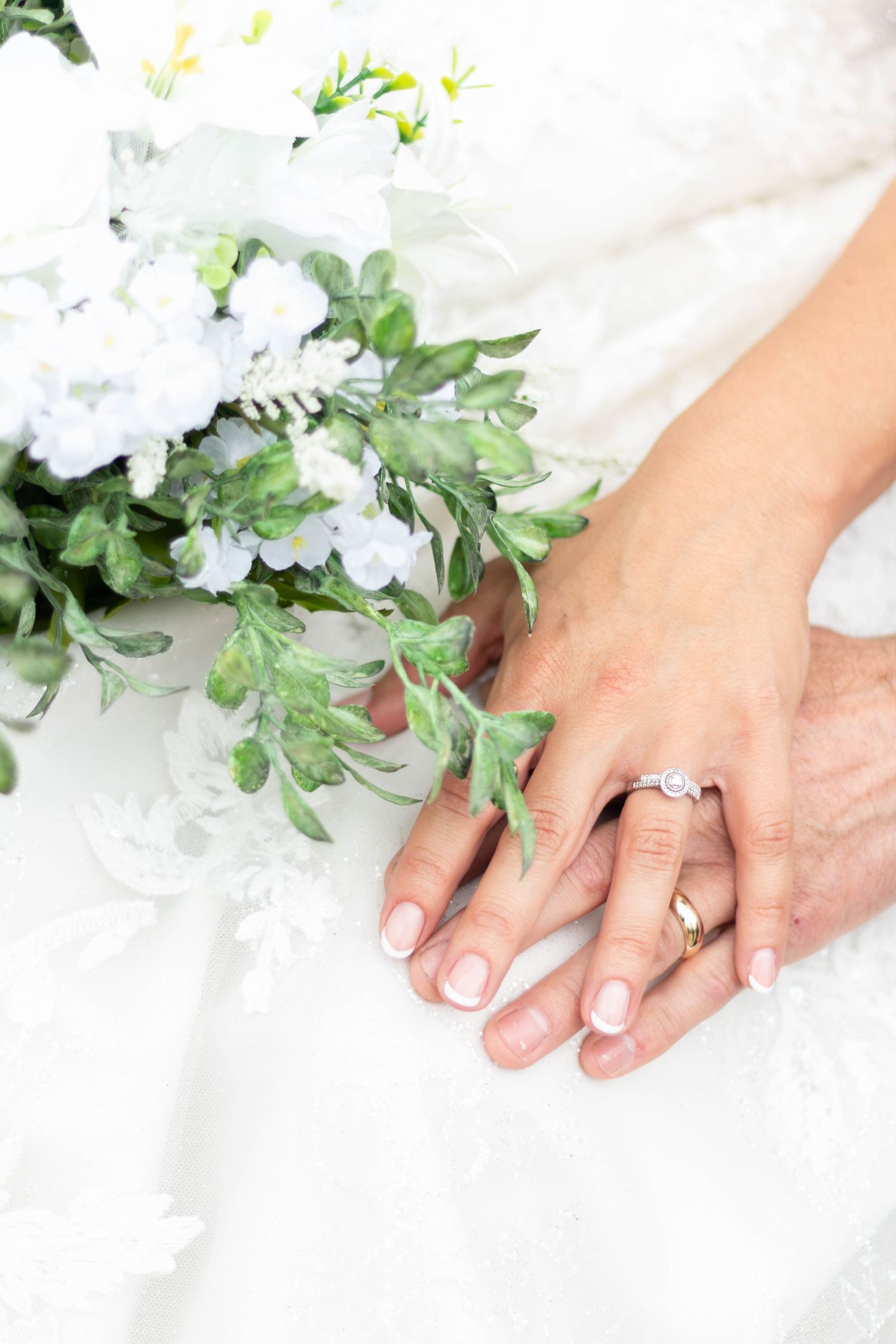 Close-up of a bride and groom holding hands beside a white and greenery wedding bouquet.