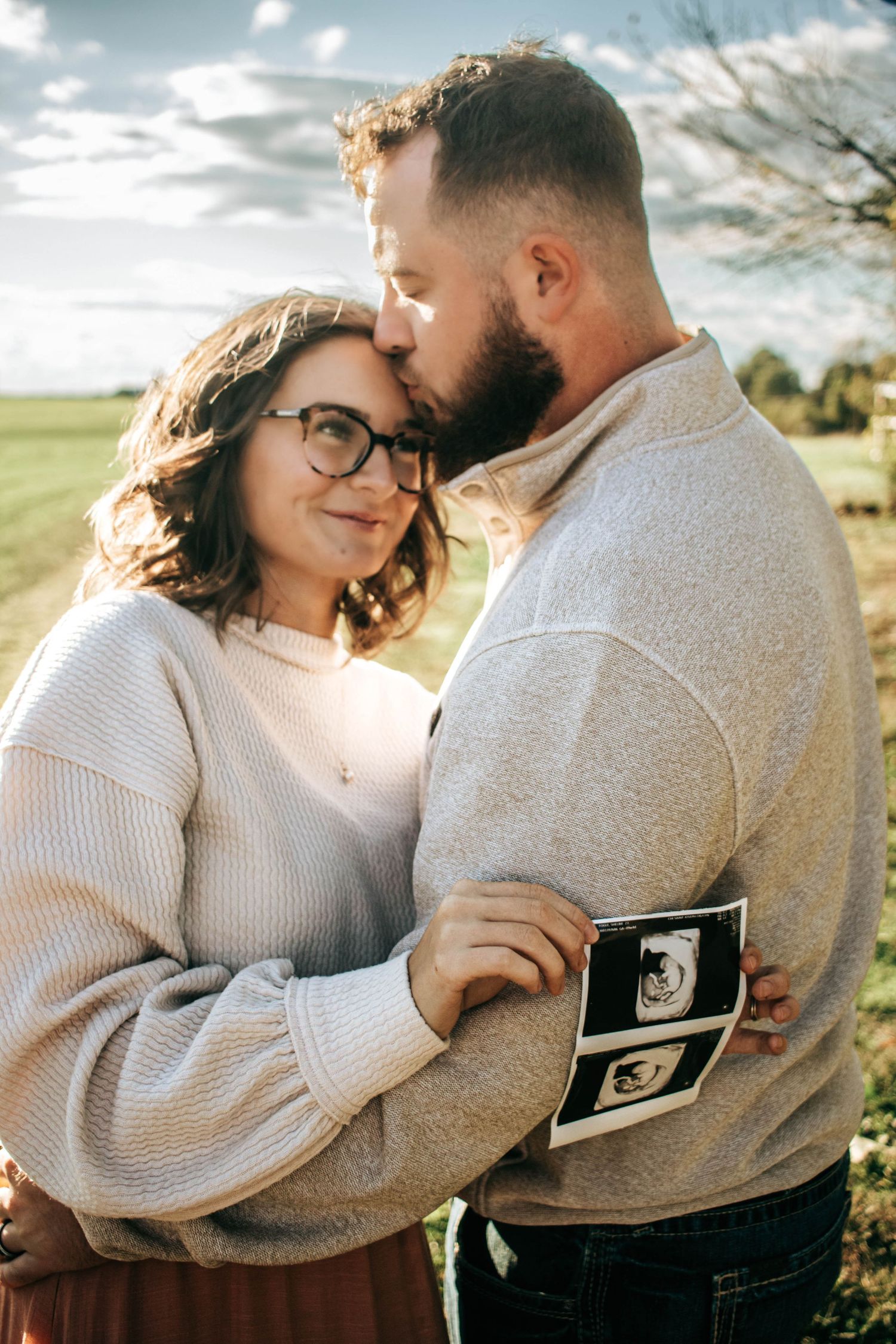 Expecting couple embracing outdoors while holding ultrasound photos.