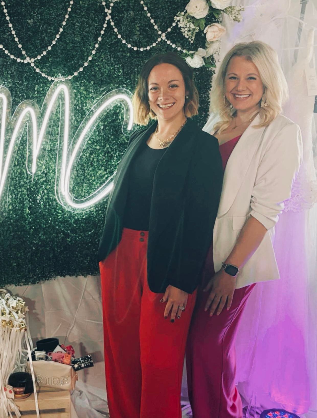 Founder Kaitlyn and manager and Kali Jo posing in front of greenery wall at bridal expo