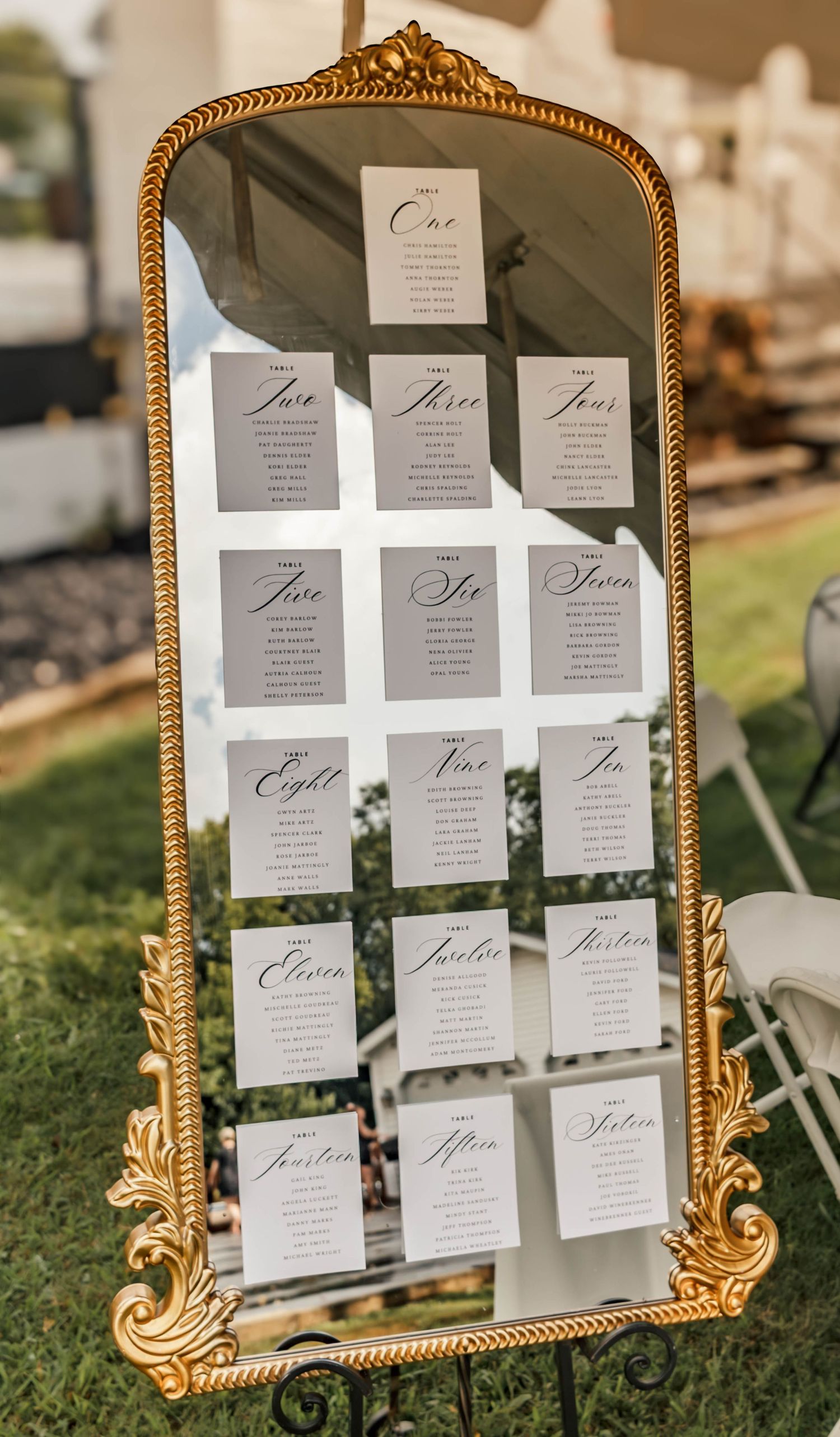 Wedding seating chart displayed on an ornate gold mirror at an outdoor wedding.
