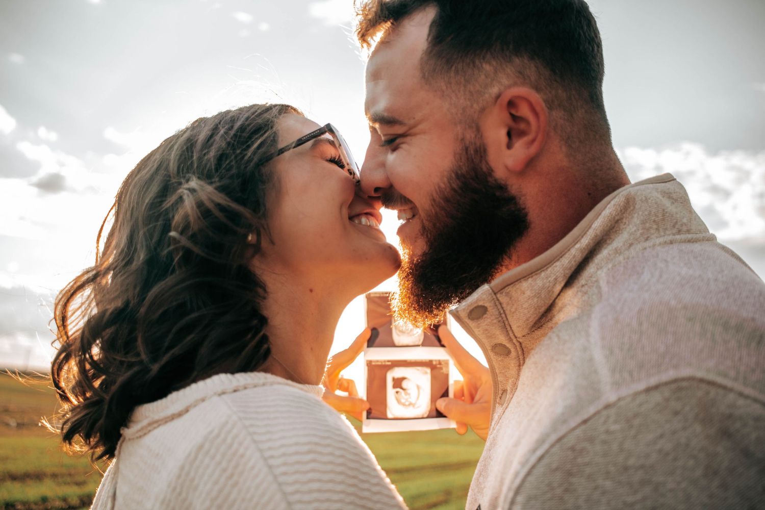 Expecting couple embracing outdoors while holding ultrasound photos.