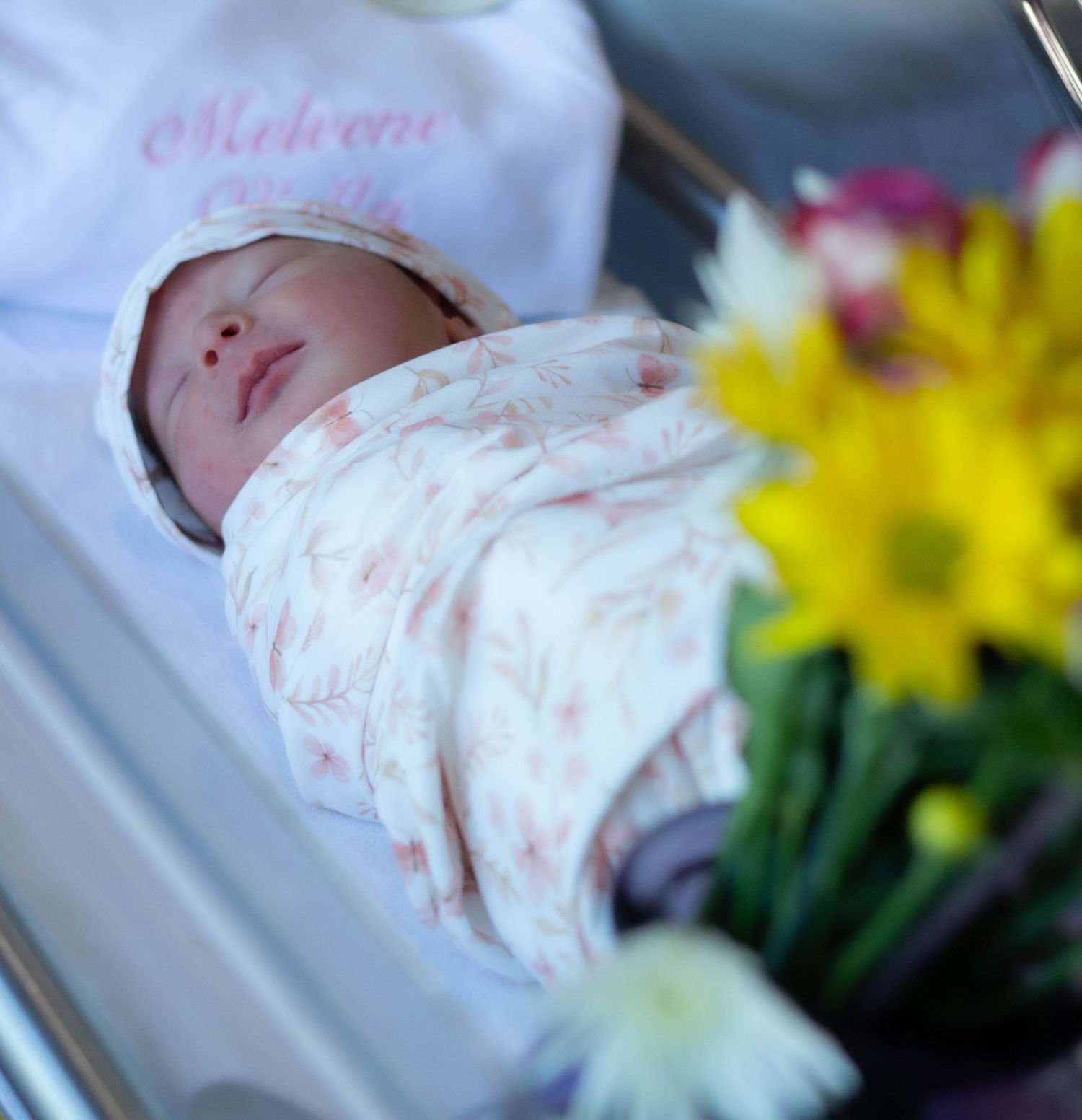 Newborn baby wrapped in a hospital bassinet beside flowers.