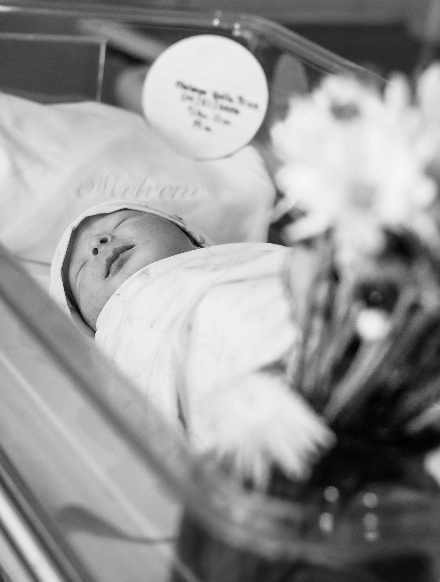 Black-and-white newborn portrait of a swaddled baby in a hospital bassinet.