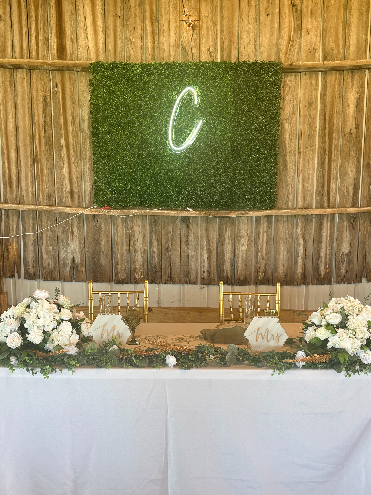 Greenery wall backdrop with illuminated initial behind a wedding sweetheart table.