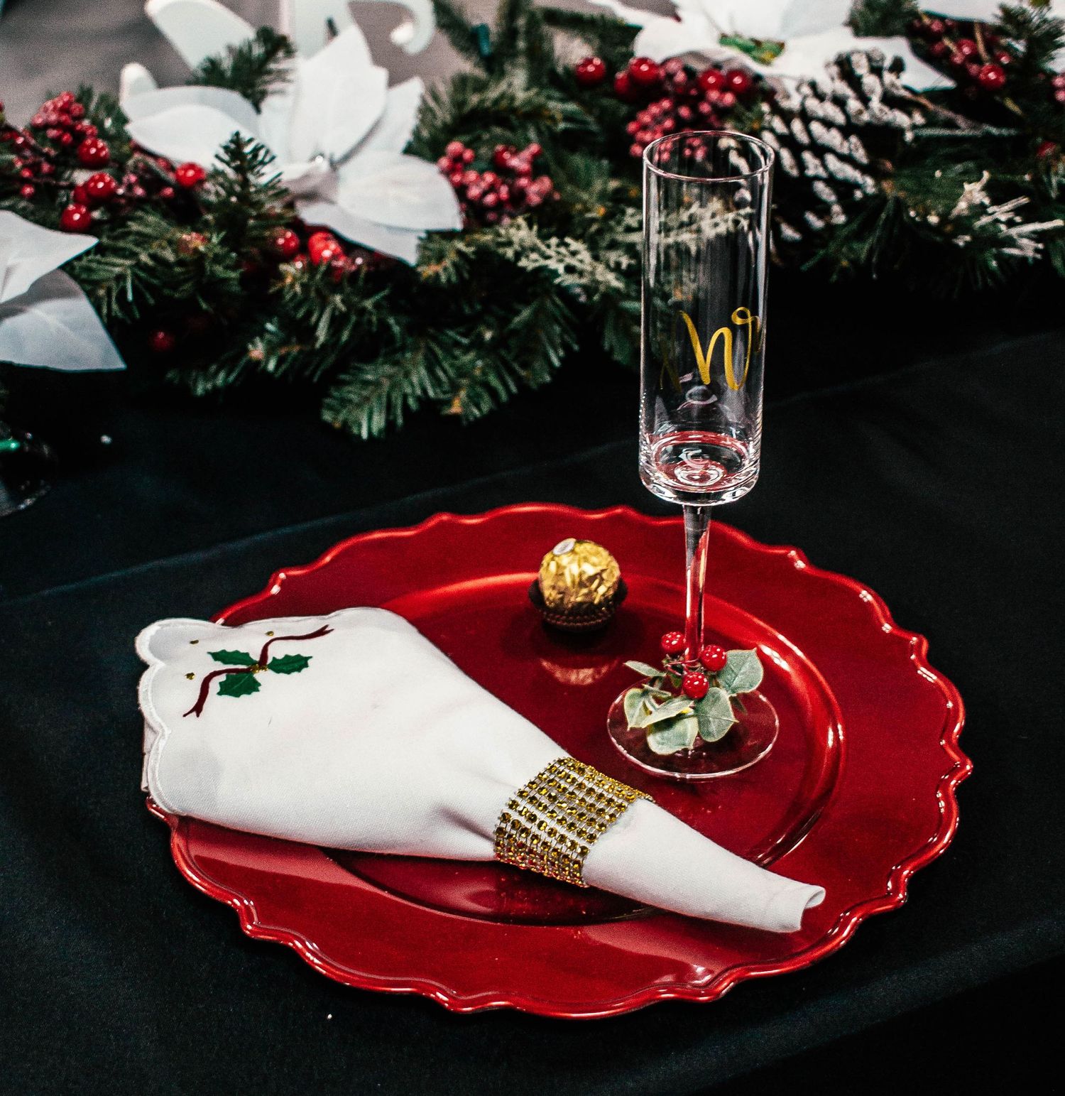 Elegant holiday event place setting with red dinnerware, white napkin, greenery, and personalized glassware.