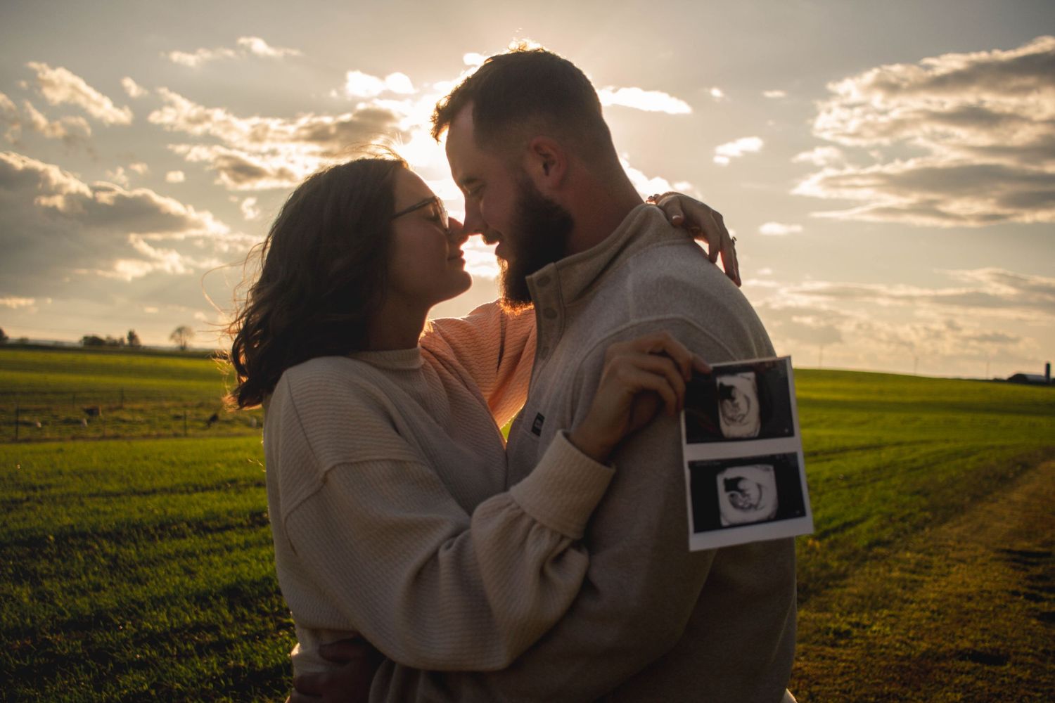 Expecting couple embracing in a field at sunset with ultrasound photos.