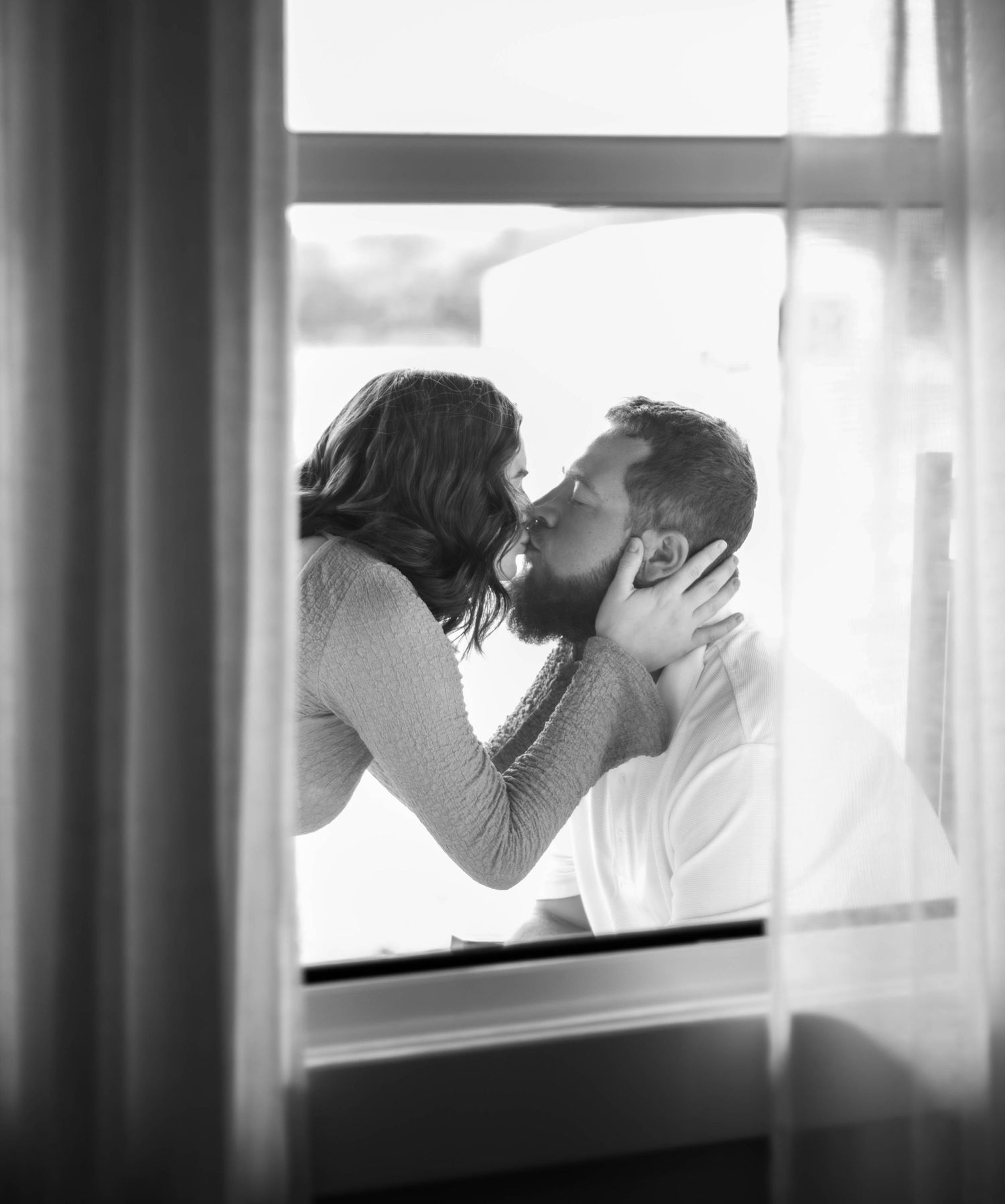 Black-and-white maternity portrait of an expecting couple kissing by a window.