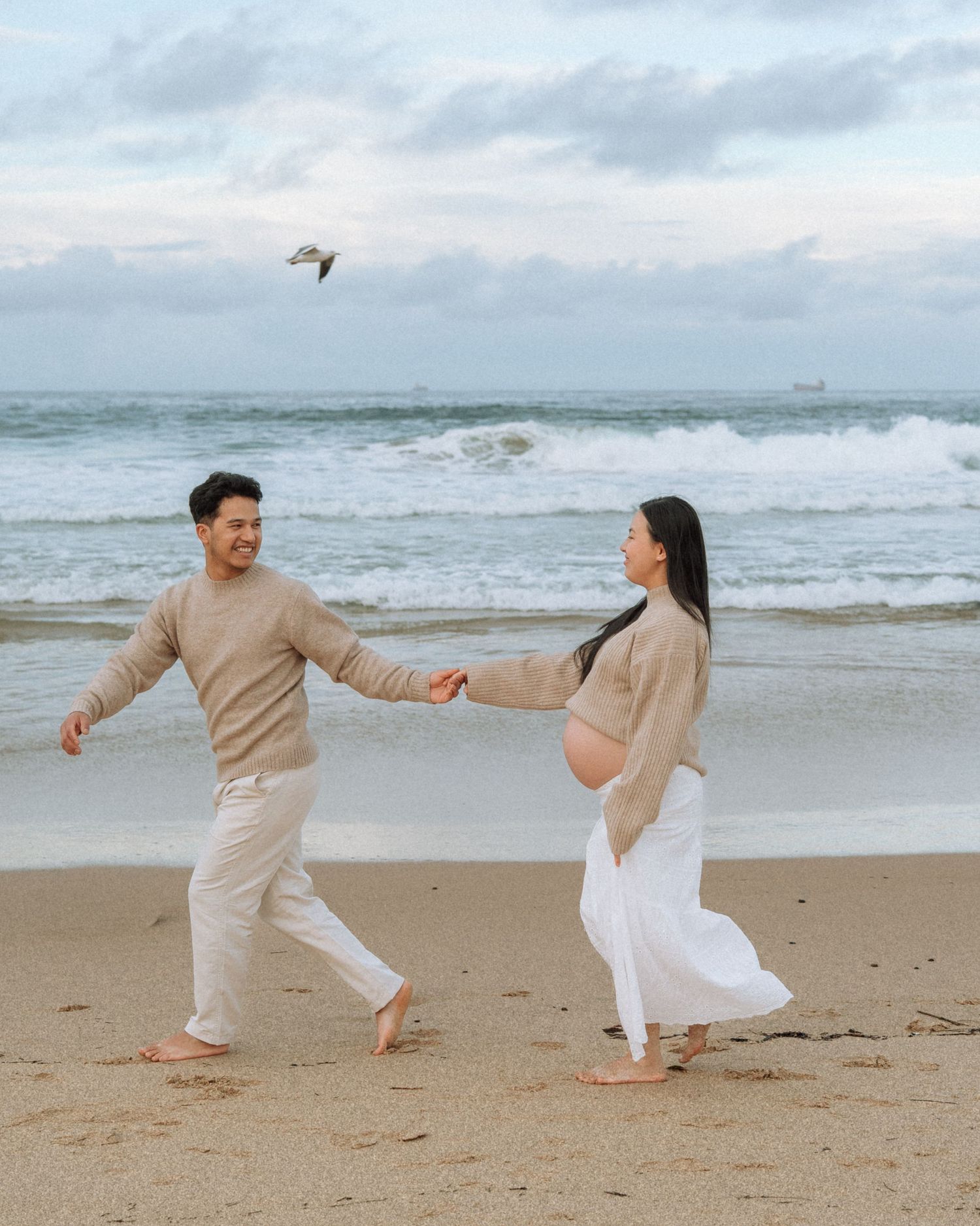 Joyful expecting couple holds hands walking along beach shoreline with crashing waves and cloudy sky behind them.