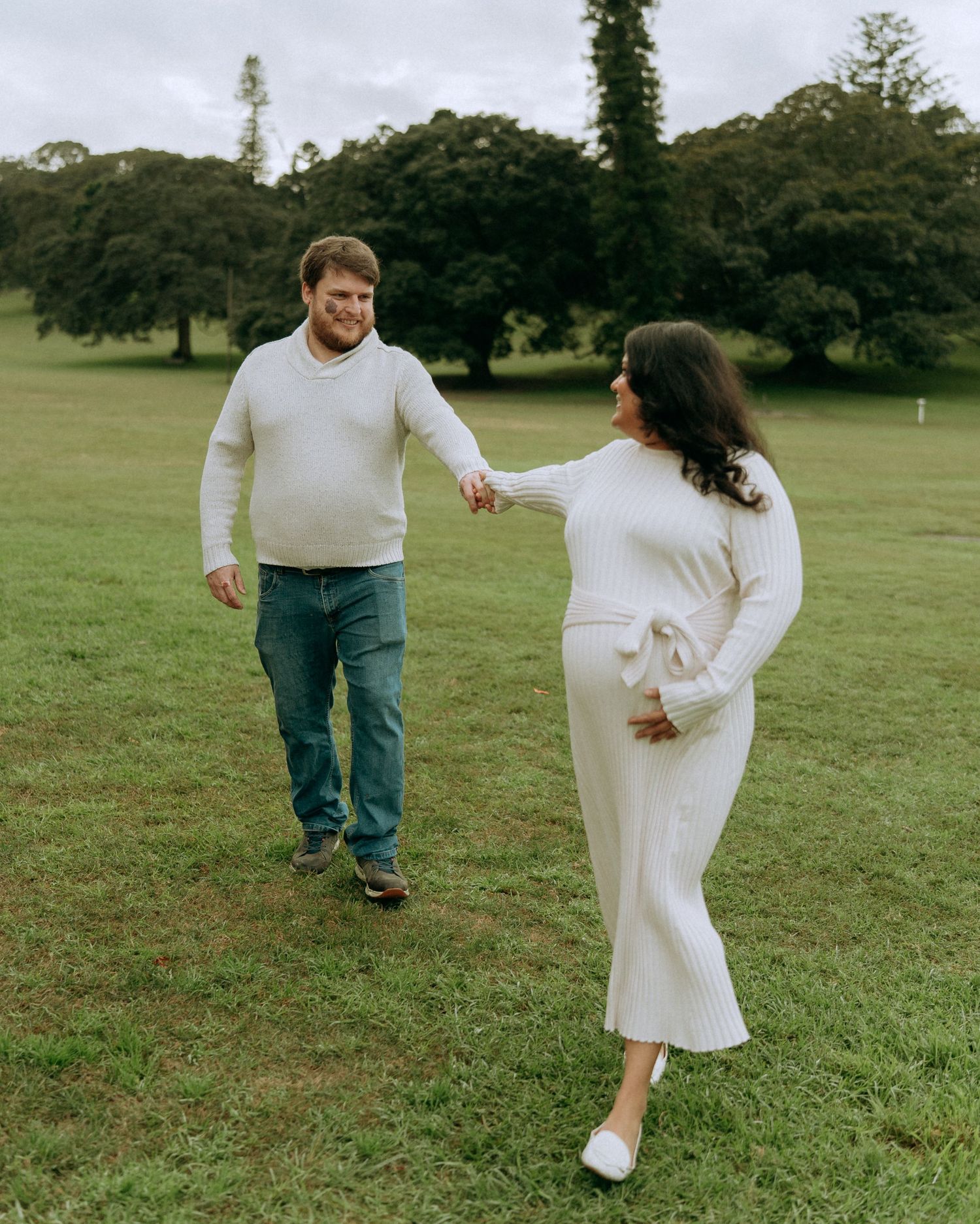 Pregnant woman in white ribbed dress dancing with partner on a green grassy field outdoors.