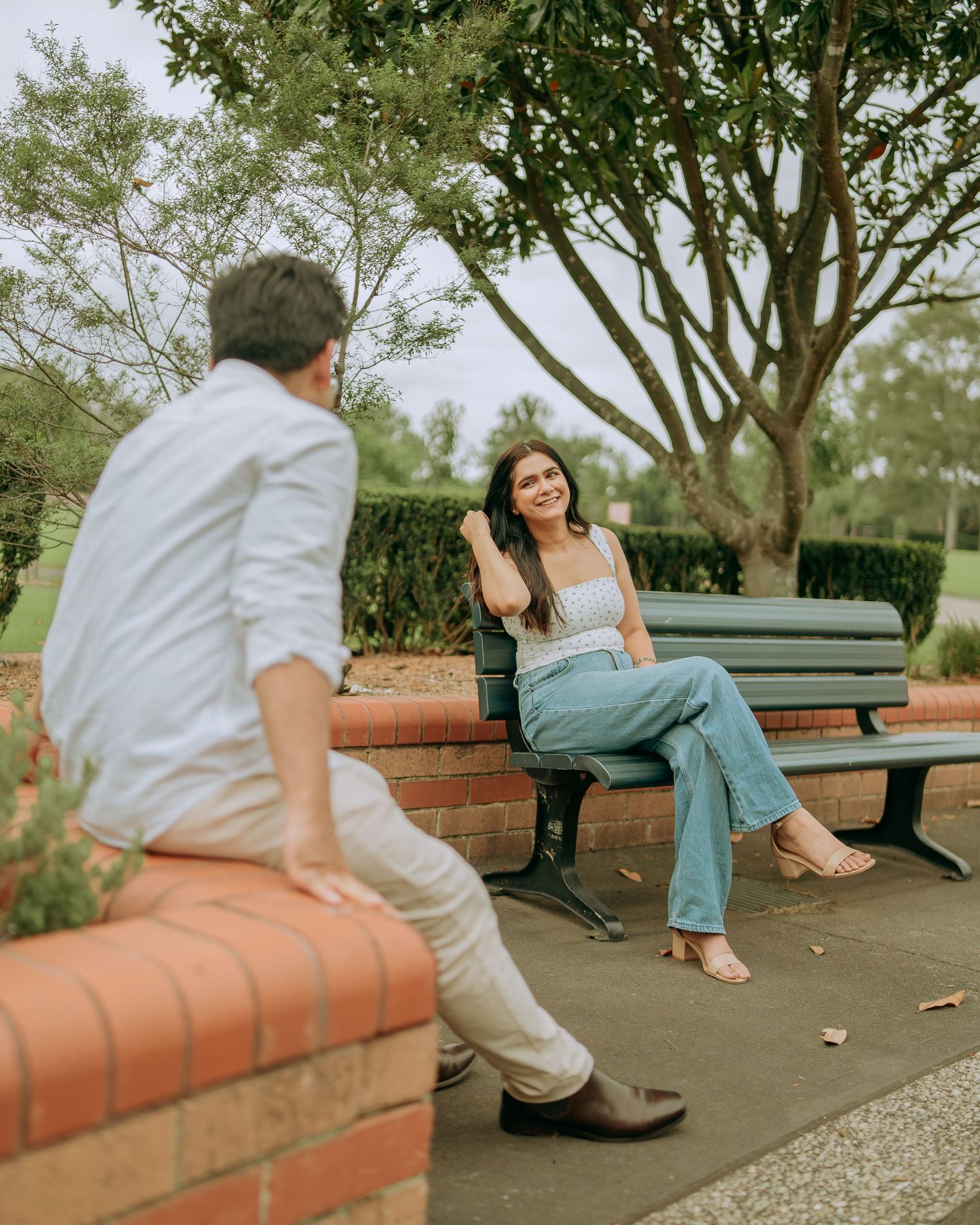 Couple sharing a smile across a brick wall and park bench surrounded by green trees.