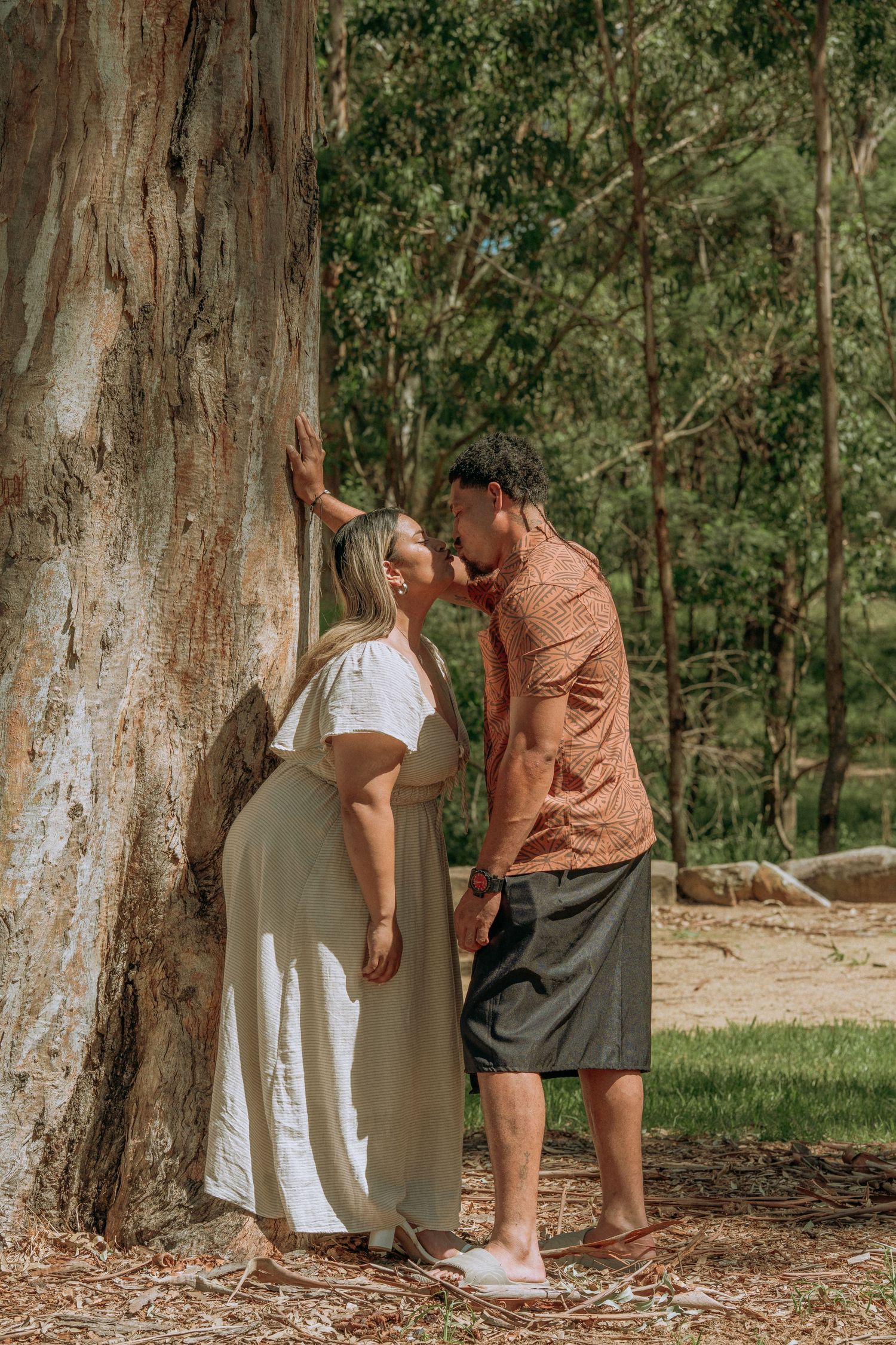 Romantic couple sharing an intimate moment leaning against a large eucalyptus tree in a lush forest setting.