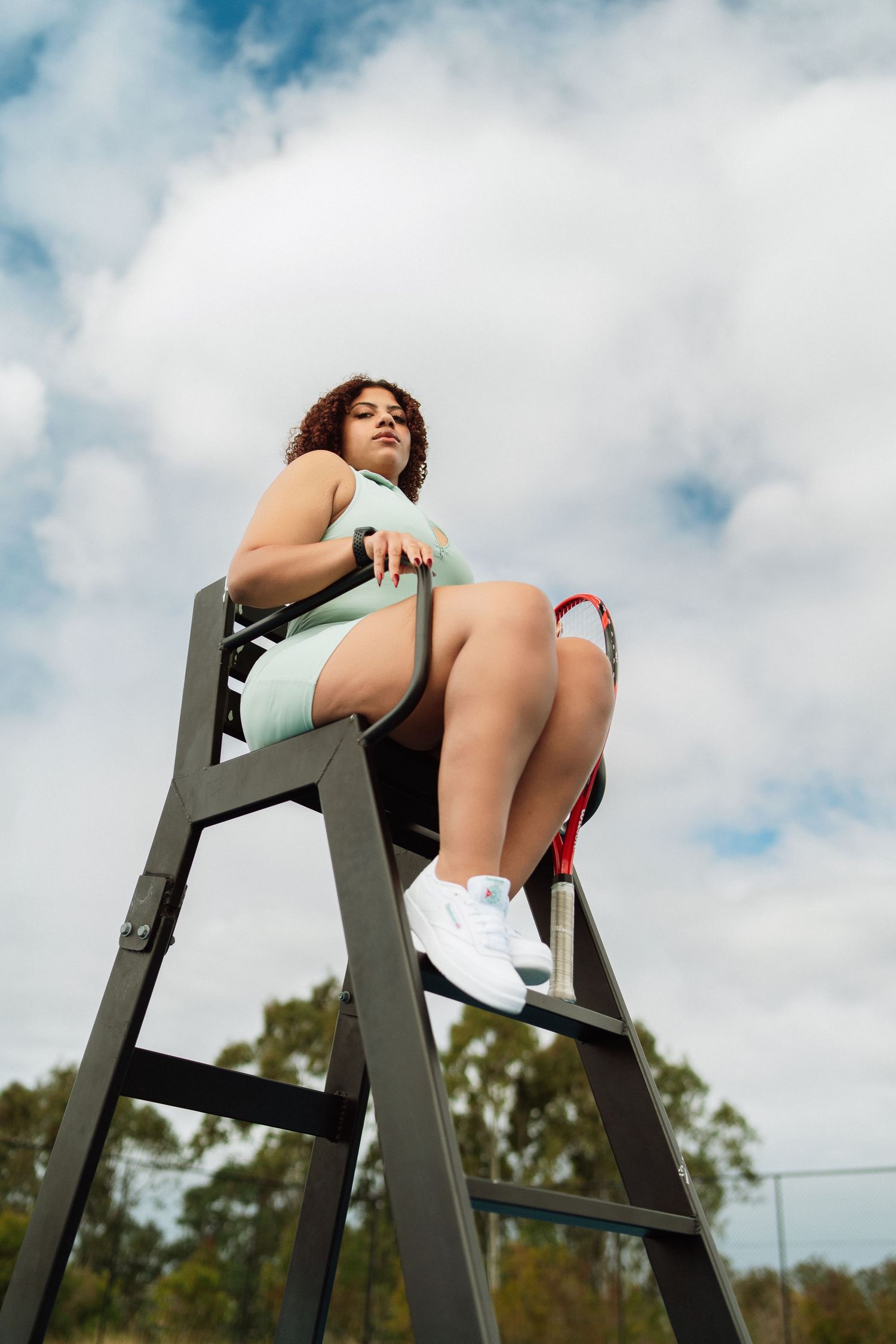A curly-haired woman in mint green tennis dress sits atop a referee chair on an outdoor court under cloudy skies.