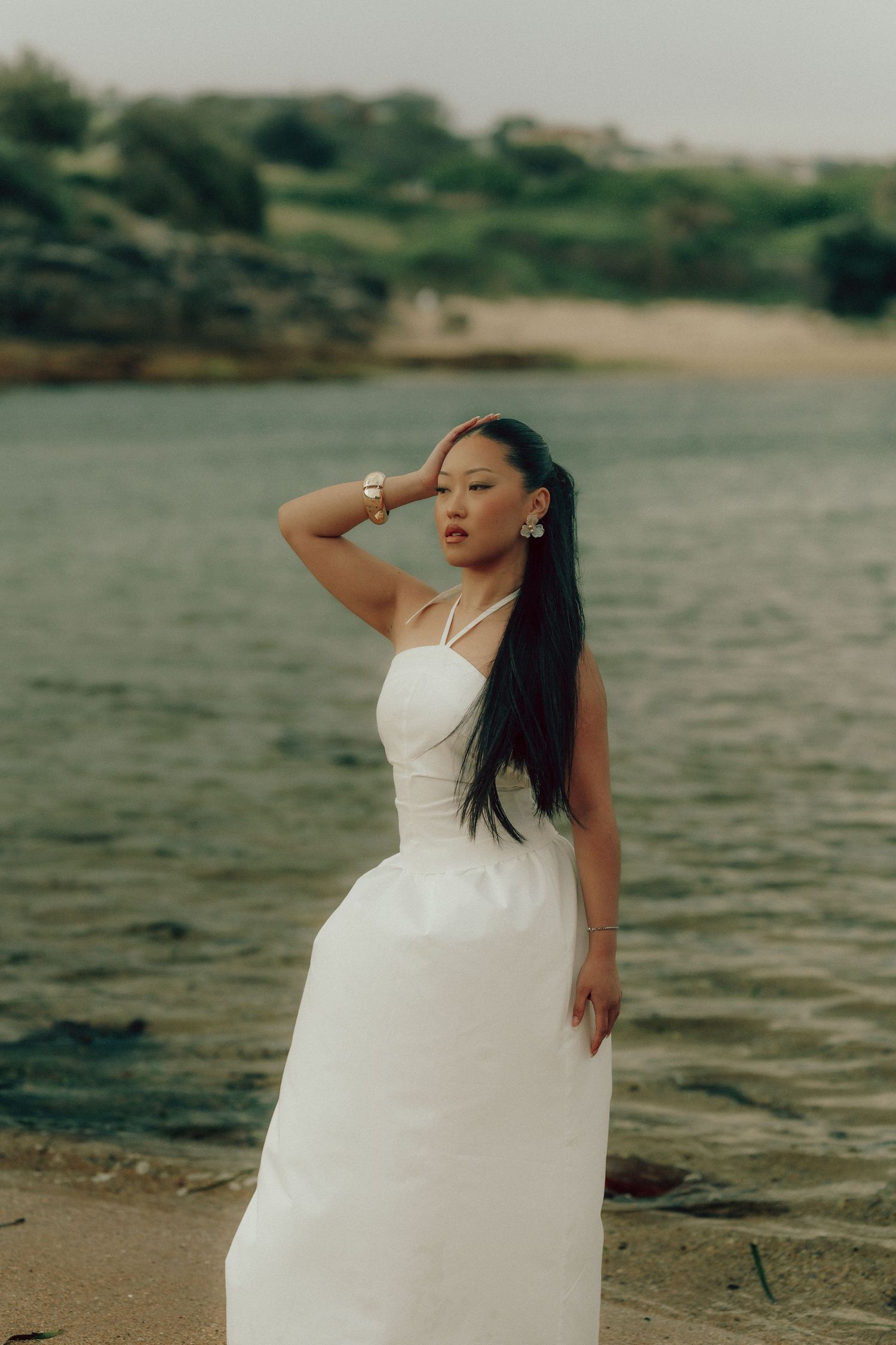 Woman in white halter dress standing on sandy shore shielding eyes with hand and green hills behind.