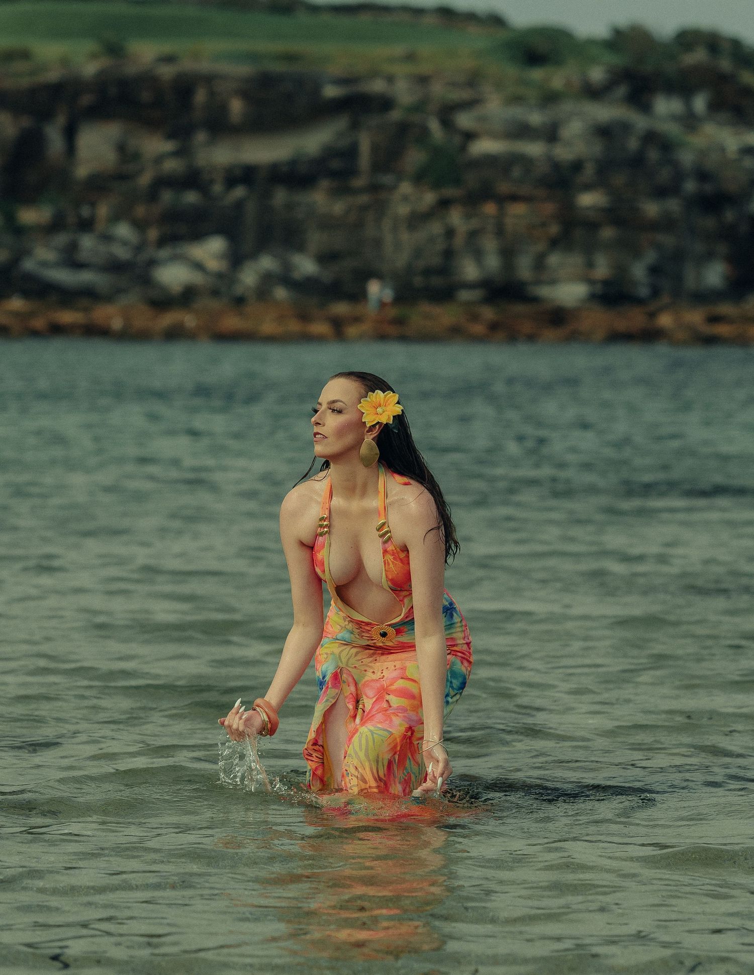 Woman in colorful floral dress wading through ocean water with yellow flower in hair at dusk.