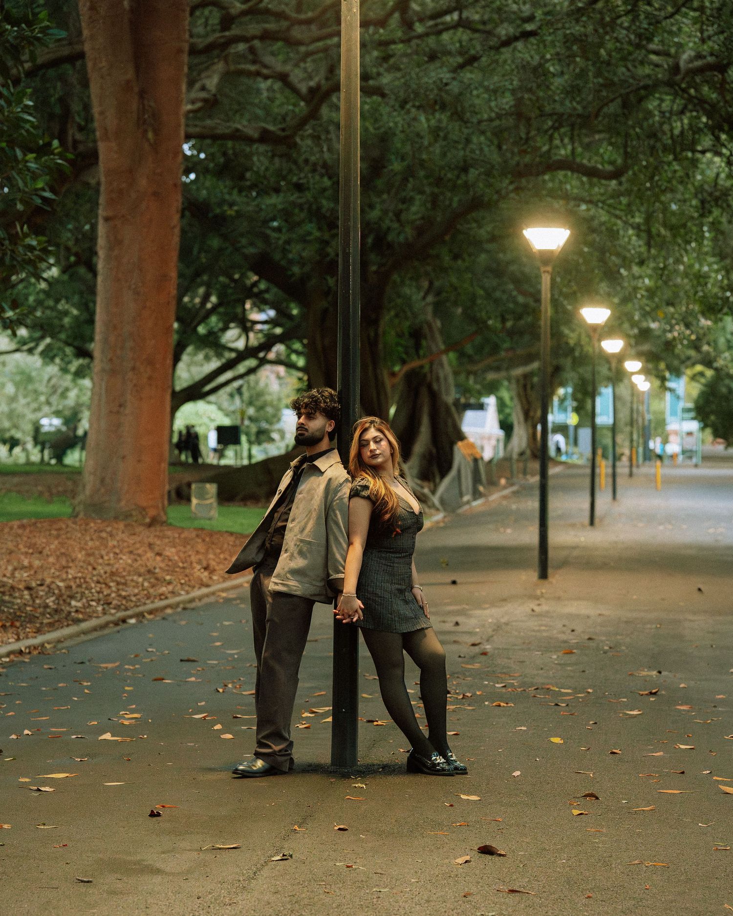 Couple leaning against a lamp post on a tree-lined park pathway at dusk.