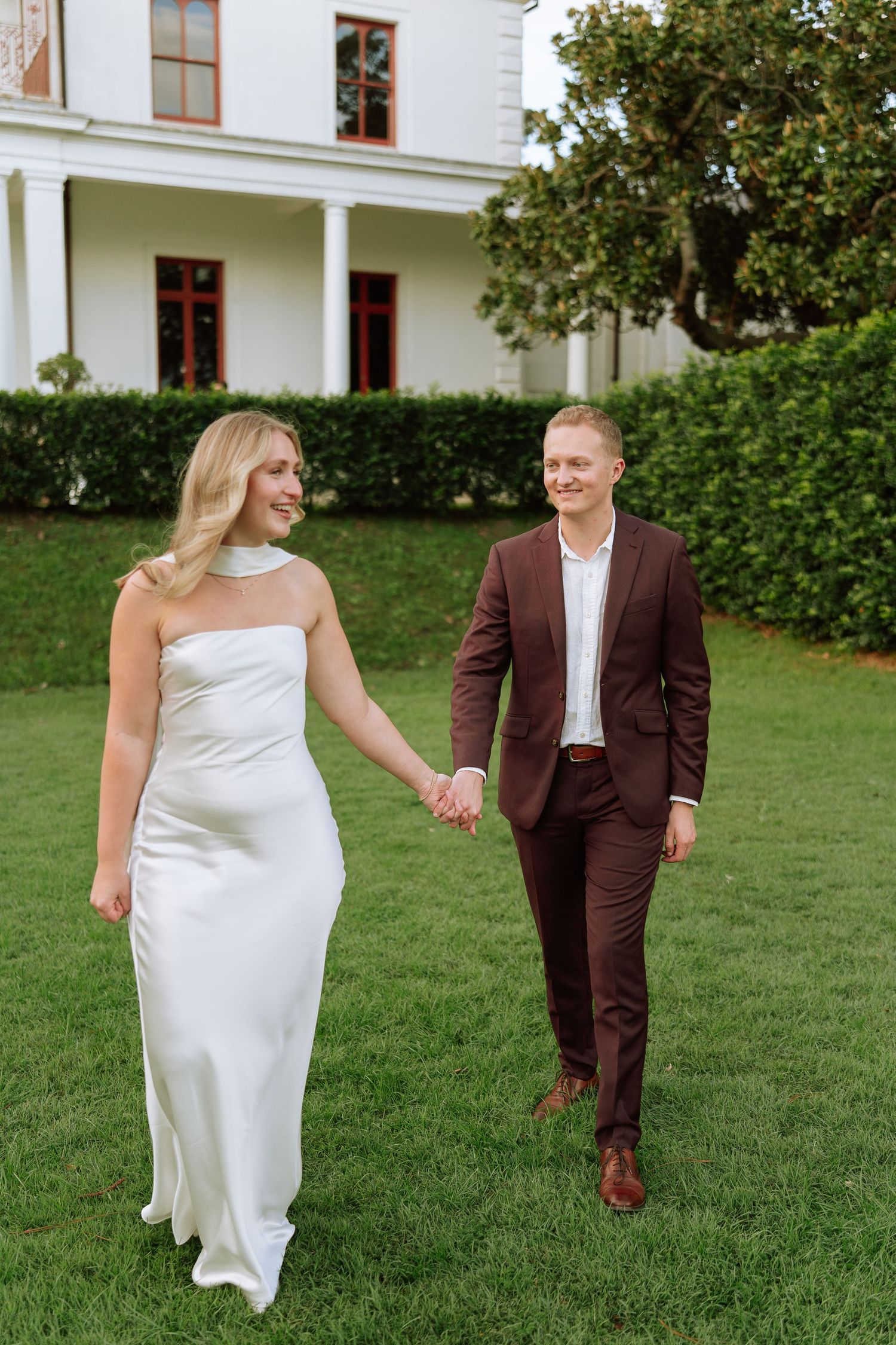 Bride in white satin gown and groom in burgundy suit walk hand in hand on green lawn near estate.