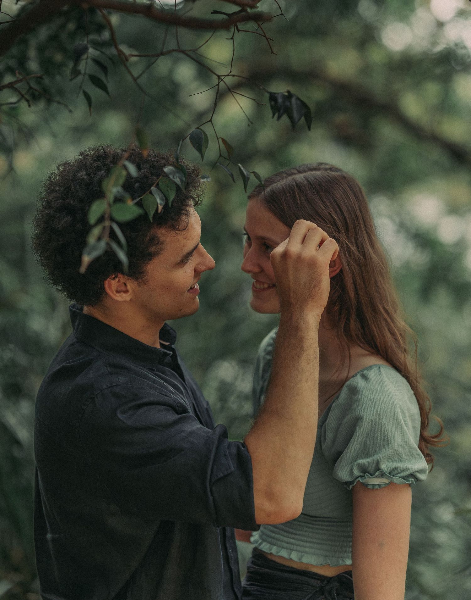 Couple smiling face-to-face under green tree branches, man gently touching womans hair.