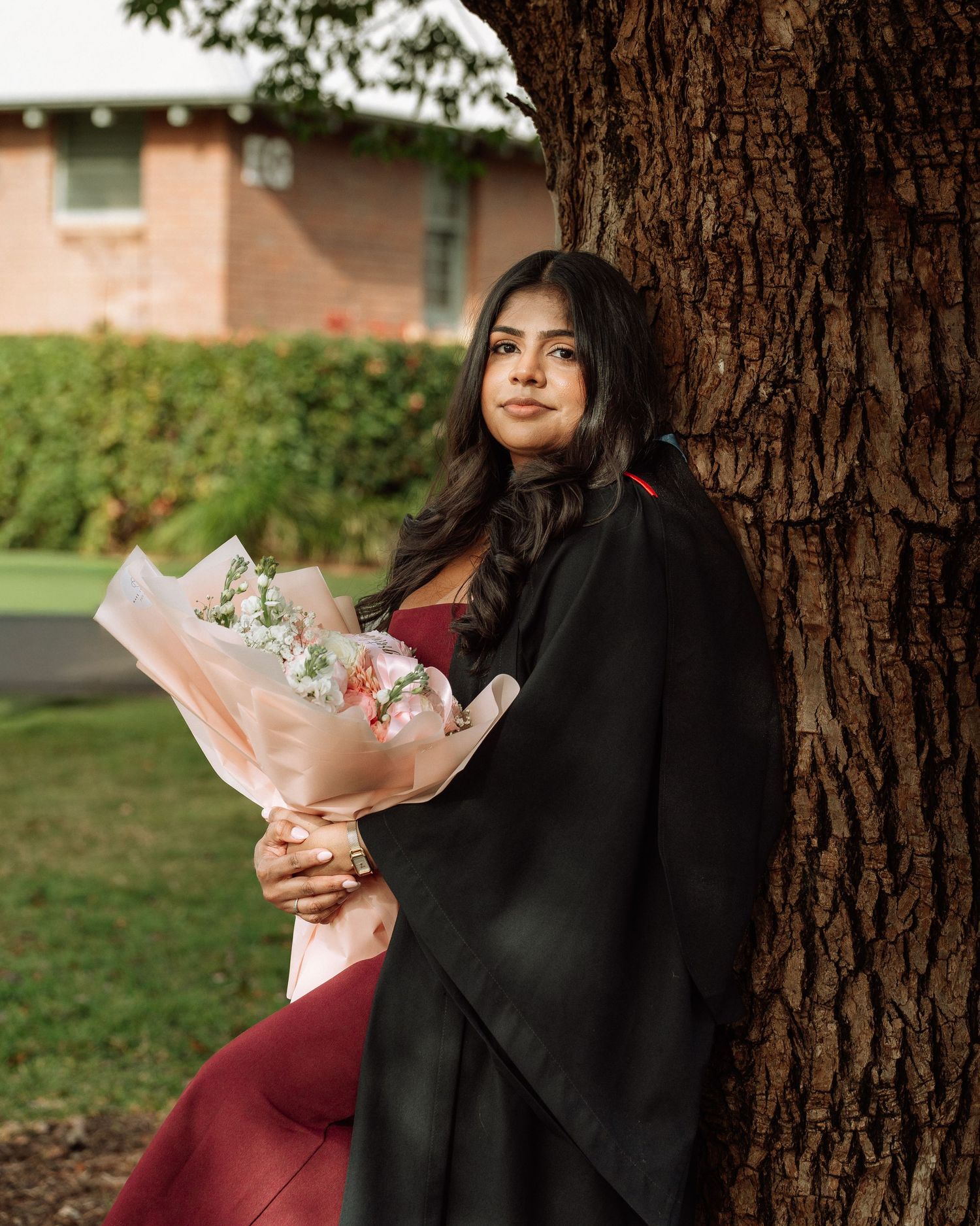 Young woman in graduation gown holds floral bouquet while leaning against tree in outdoor setting.