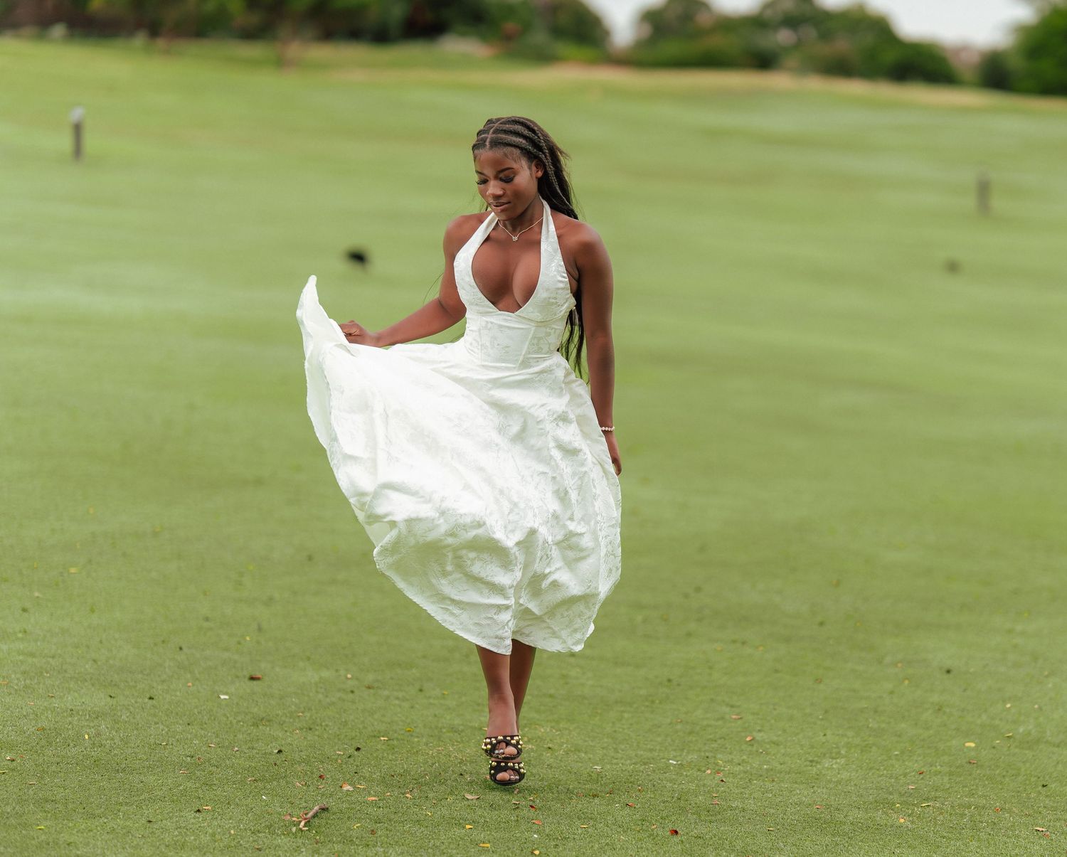 Young Black woman in flowing white halter dress twirls on a green golf course in heeled sandals.