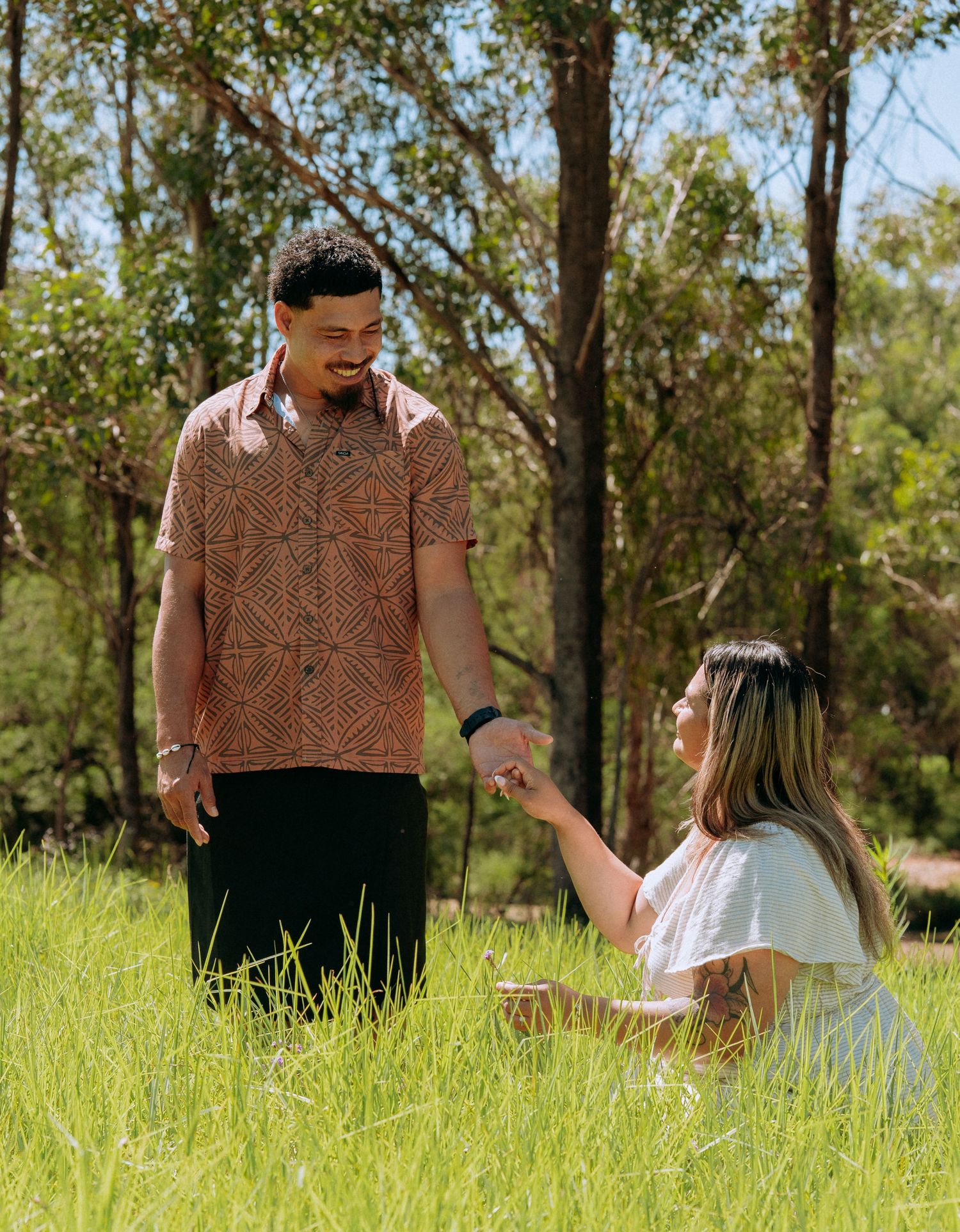 Man smiling down at woman sitting in tall green grass while holding hands in a sunny outdoor park.