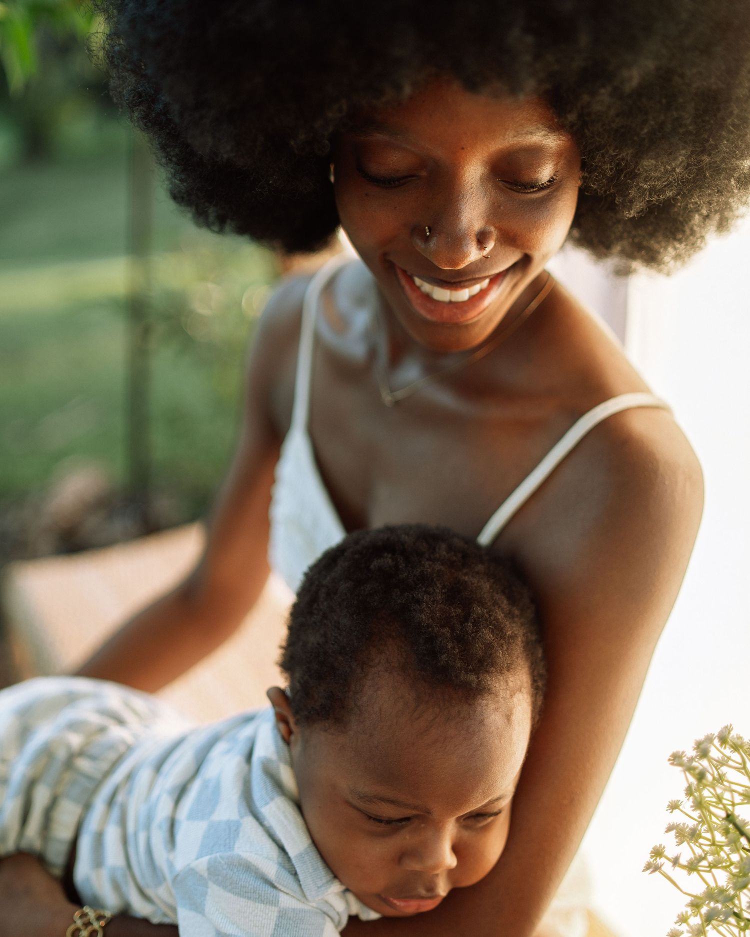 Smiling Black mother with afro hairstyle lovingly holding her baby boy outdoors in warm sunlight.