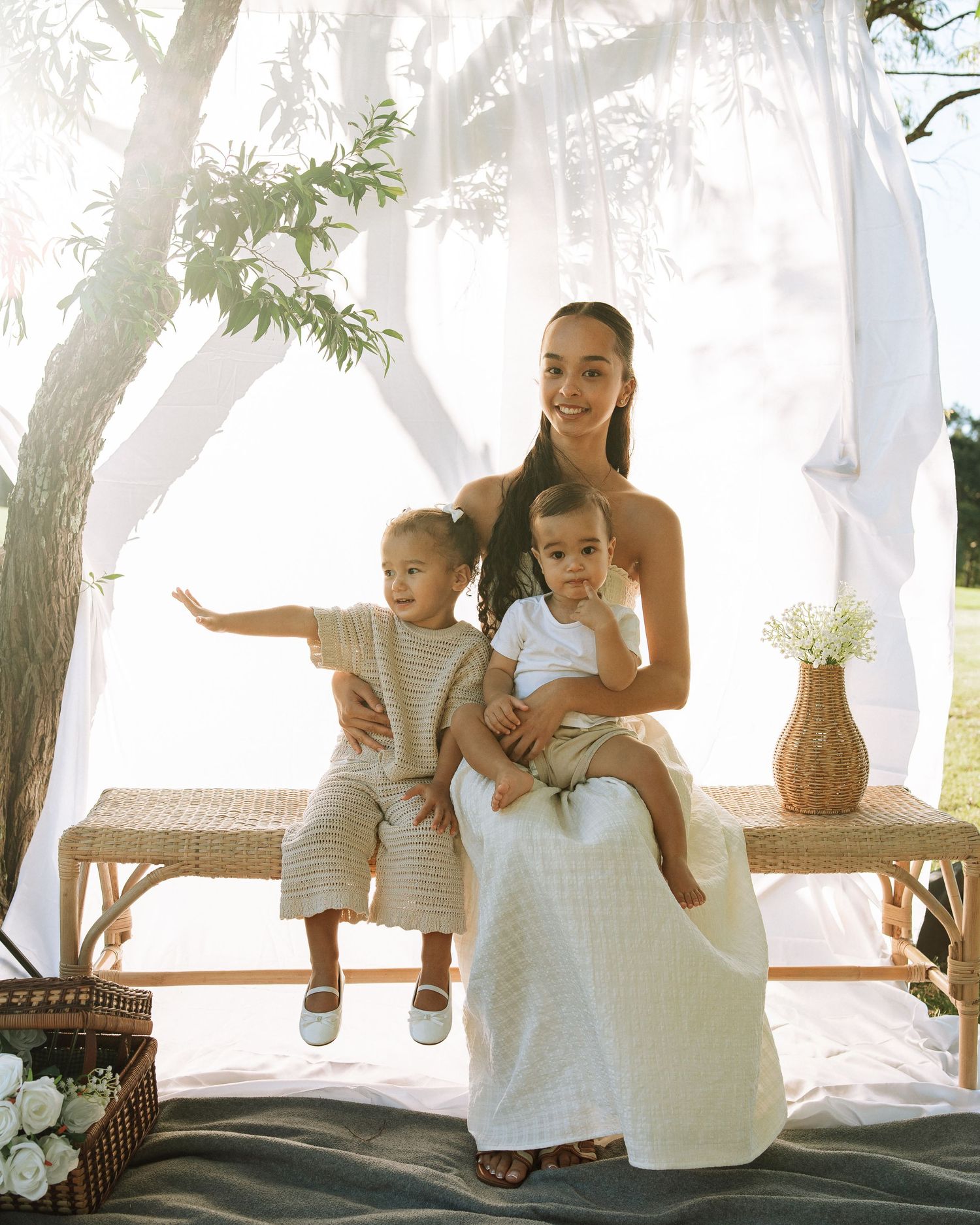 Elegant mother in white dress seated with two young children under draped canopy in styled outdoor family portrait session.