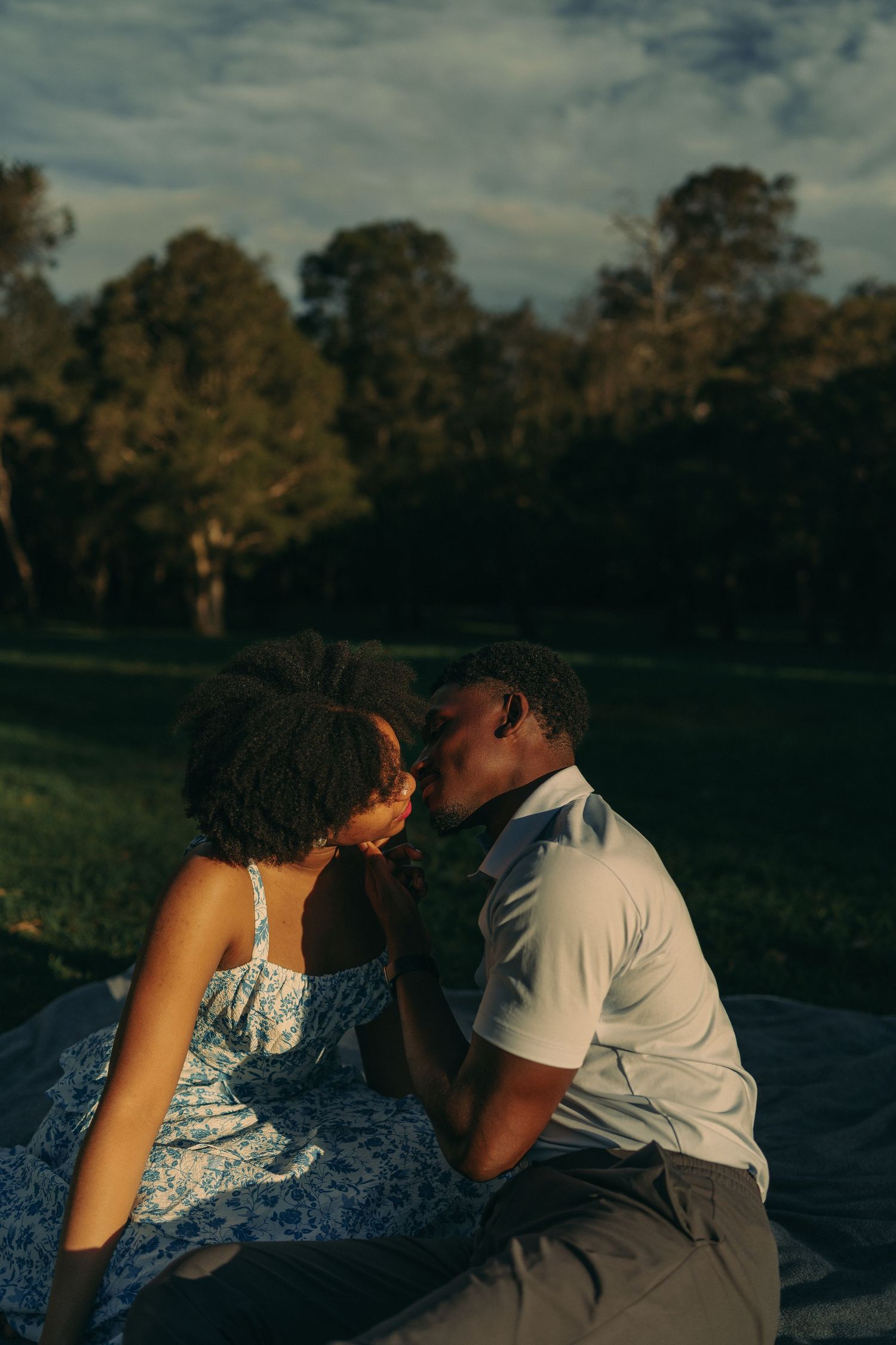 A romantic couple shares a tender kiss while sitting on a blanket in a park at sunset.
