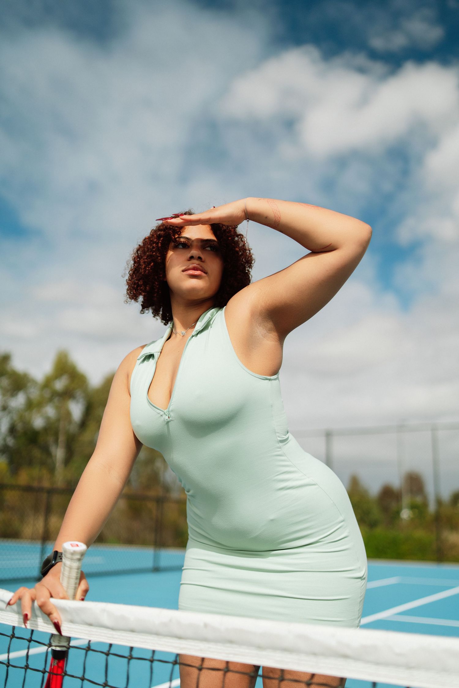 A woman in a mint green tennis dress shields her eyes from the sun while standing at the net on a blue court.