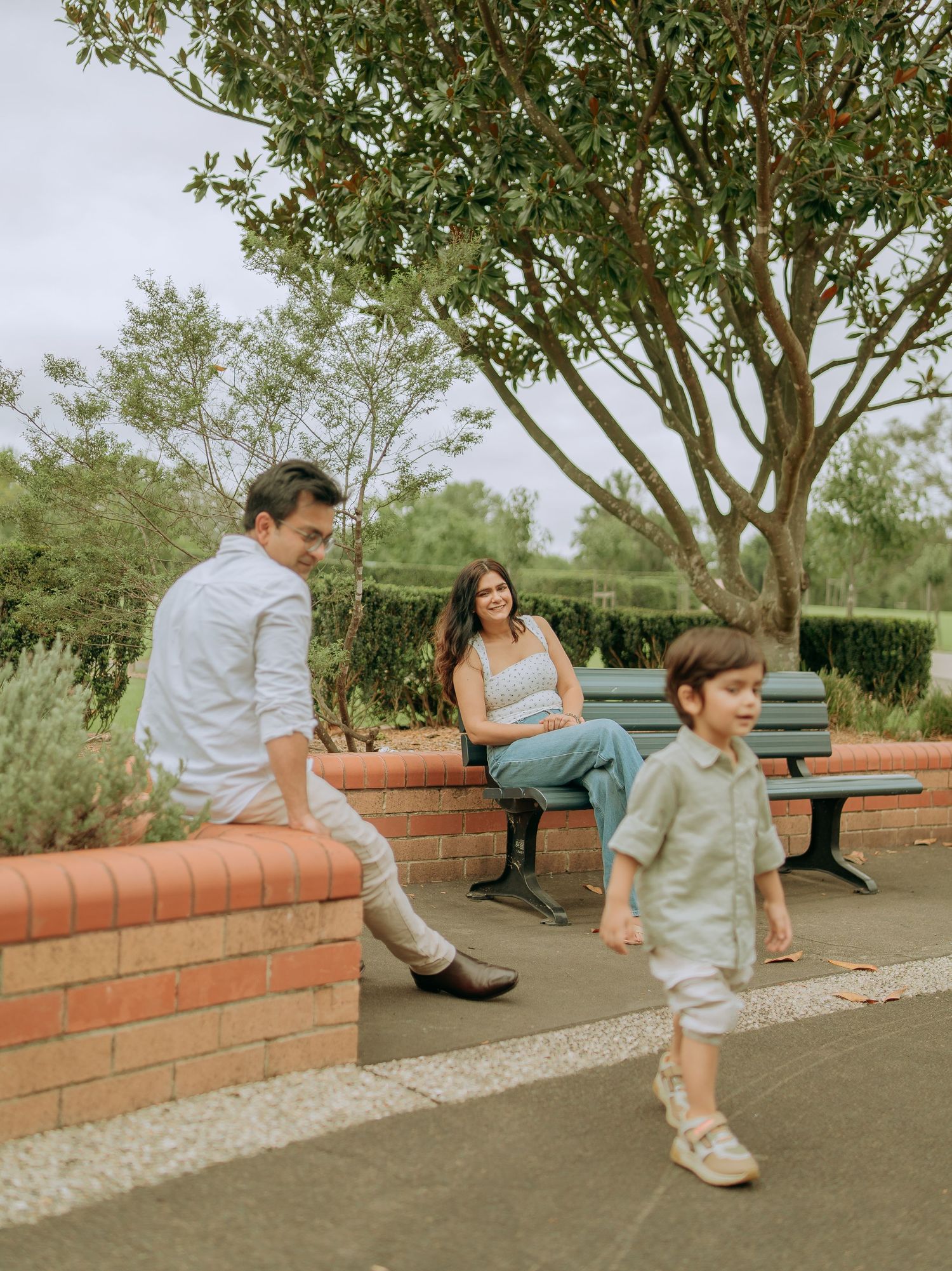Happy family in a park with parents watching their young son walk along a paved path near a bench.