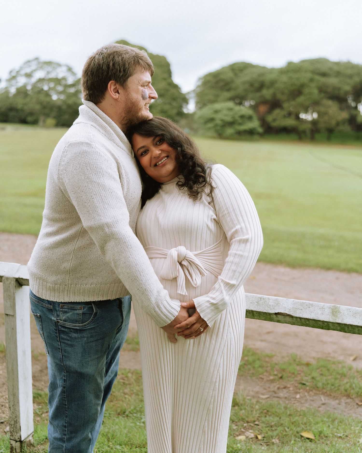 Couple embracing warmly by a white fence during an outdoor maternity photo session in a park.