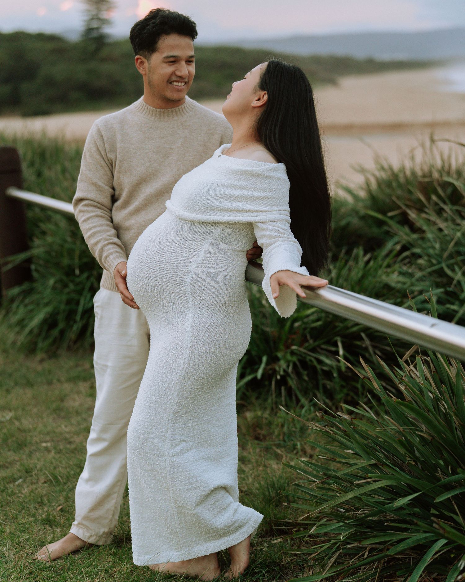 Expecting couple smiles at each other near coastal railing, woman in fitted white knit maternity dress at dusk.