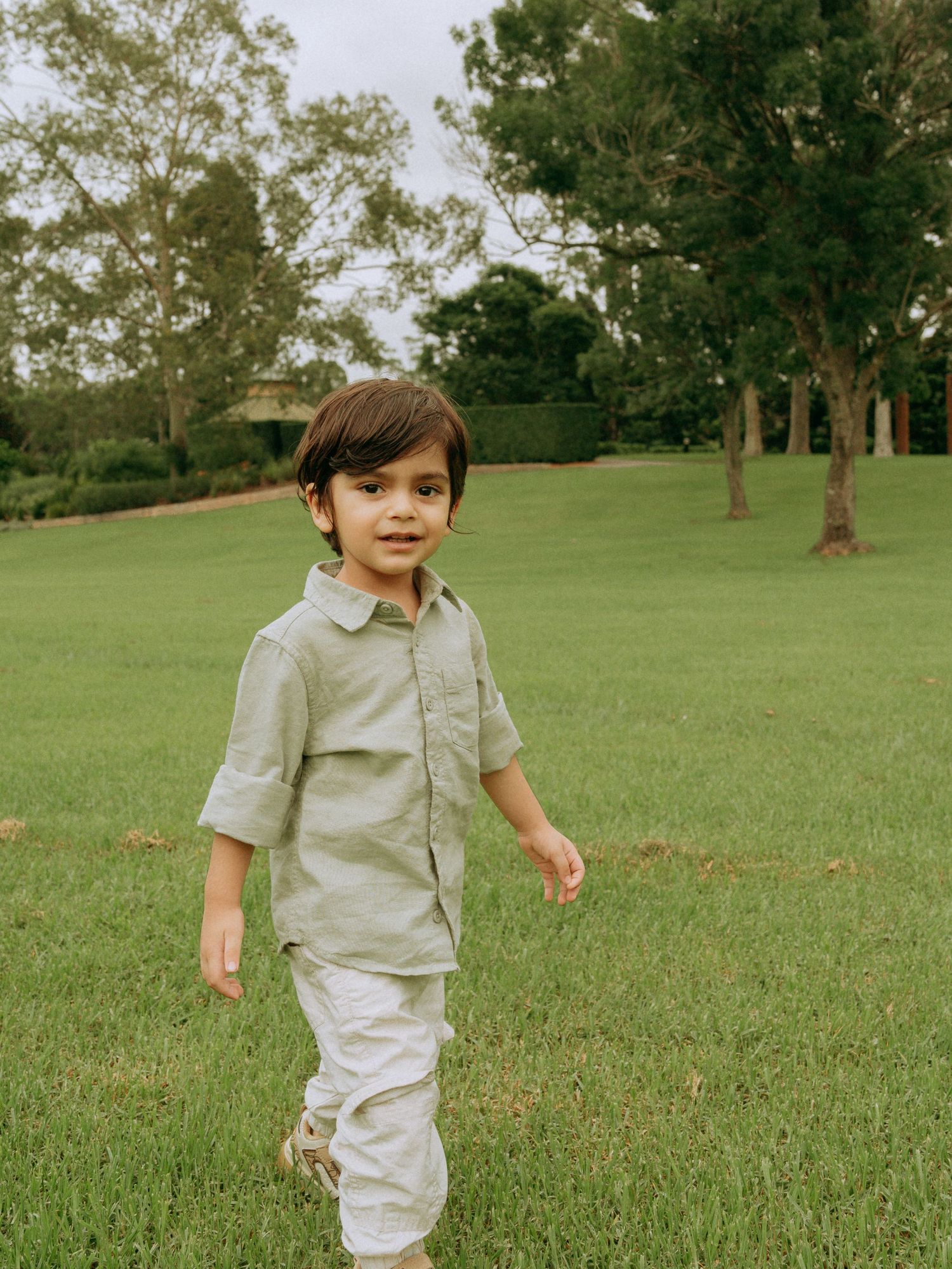 Adorable toddler boy in sage green linen shirt walking confidently across a lush green lawn.
