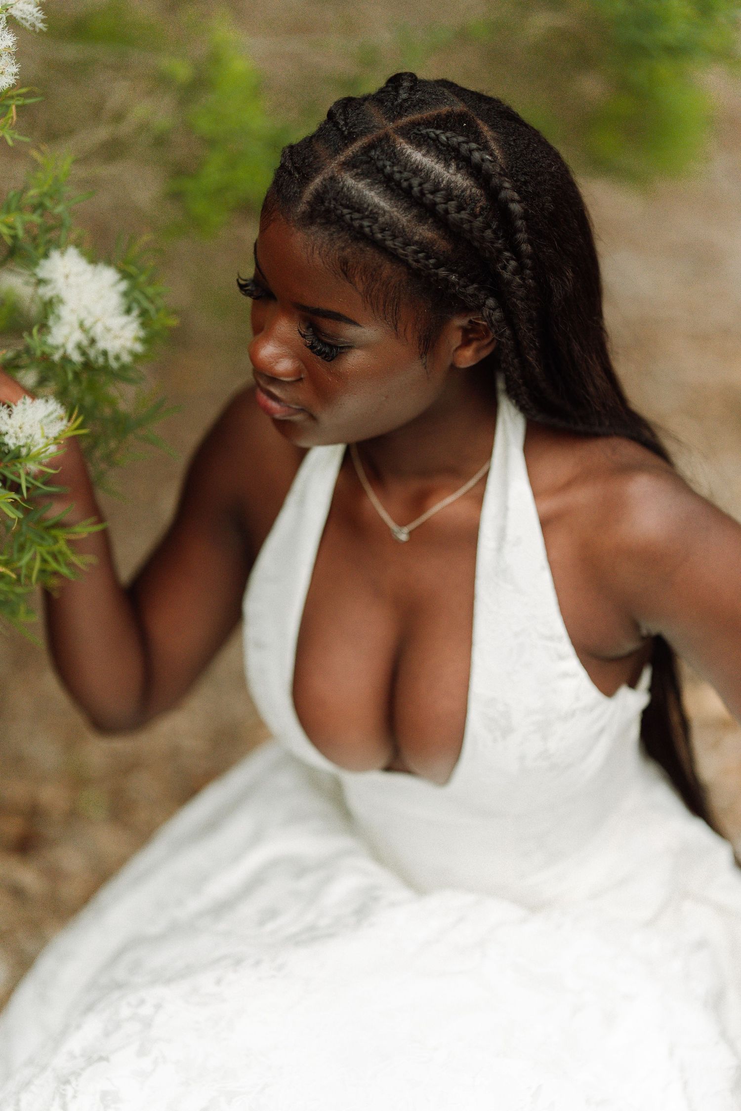 Young Black woman in white halter dress with braided hair reaches for white flowers outdoors.