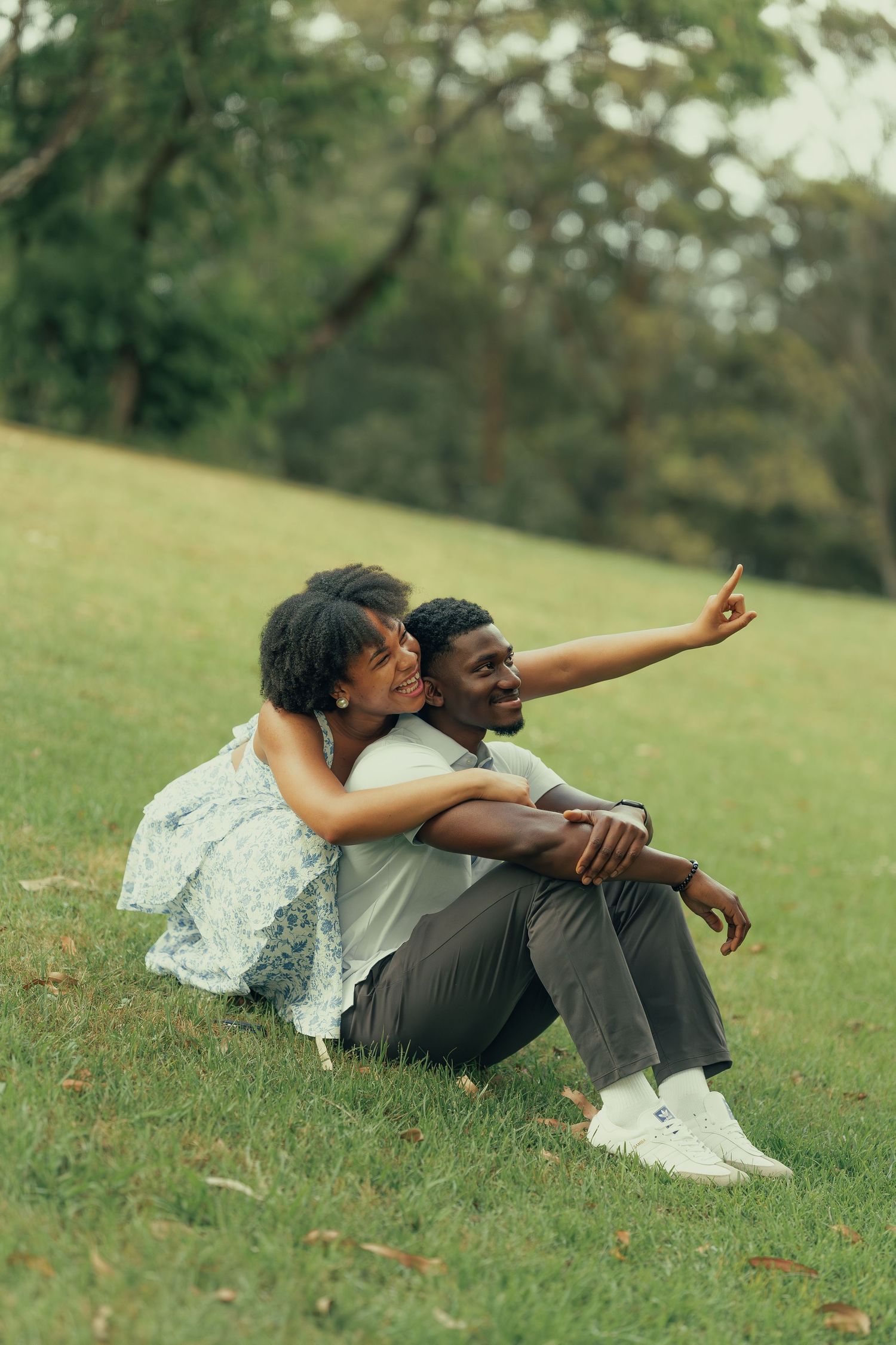 Couple sitting on grass in a park, woman embracing man as he points into the distance.