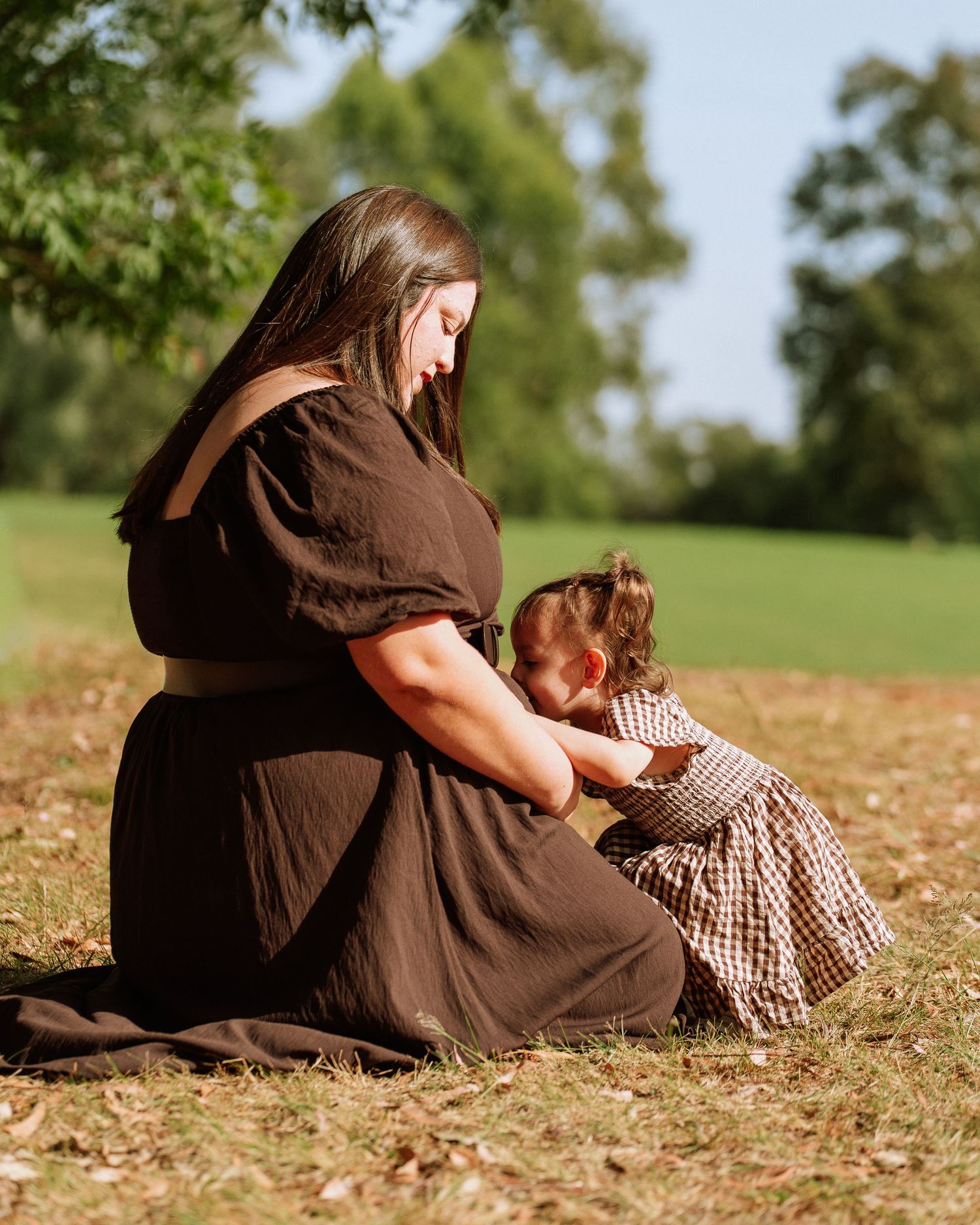 Mother in black dress sits on grass with toddler daughter in a tender outdoor maternity portrait moment.