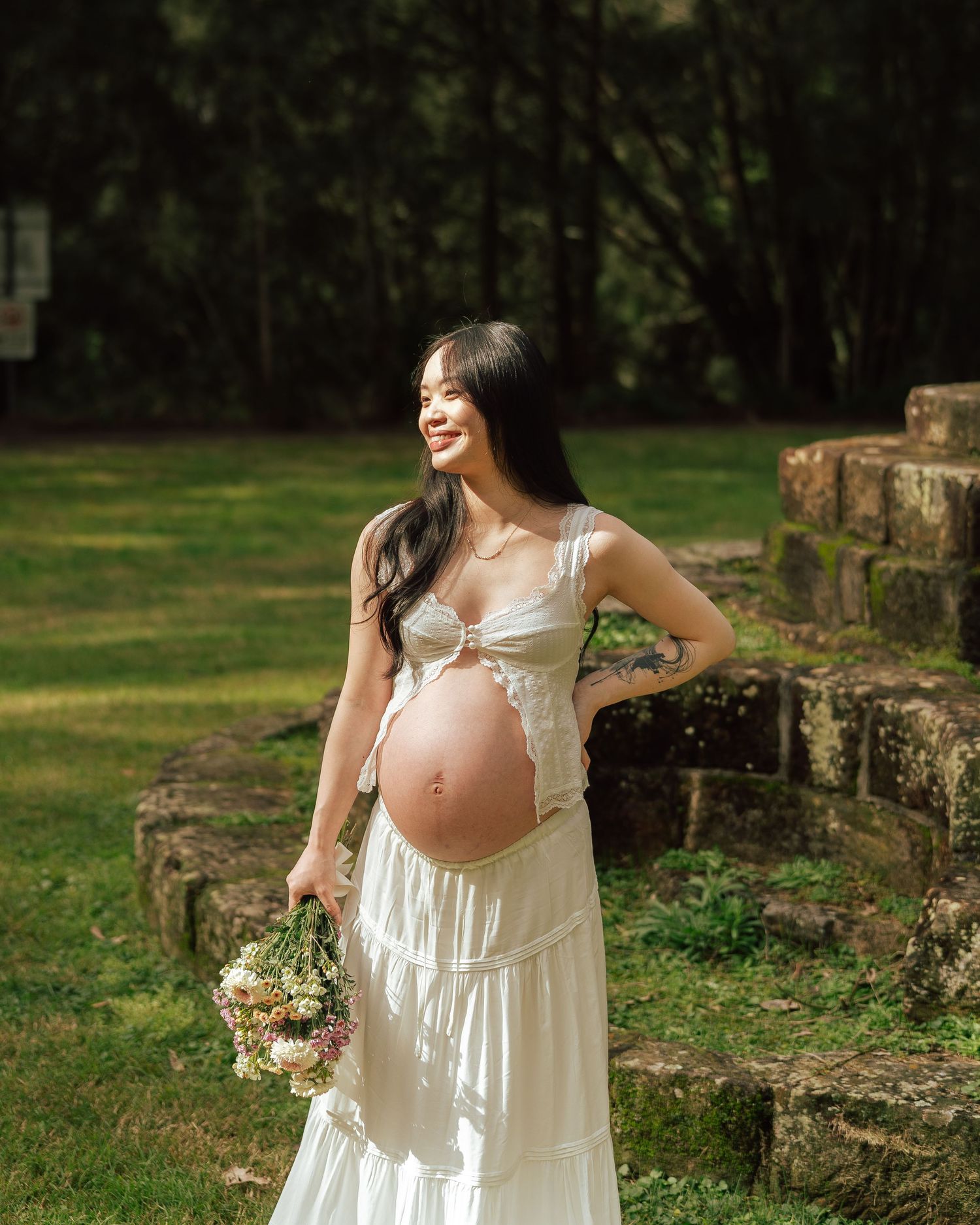 Pregnant woman in white dress holding flowers, smiling outdoors near stone steps in a lush green park.