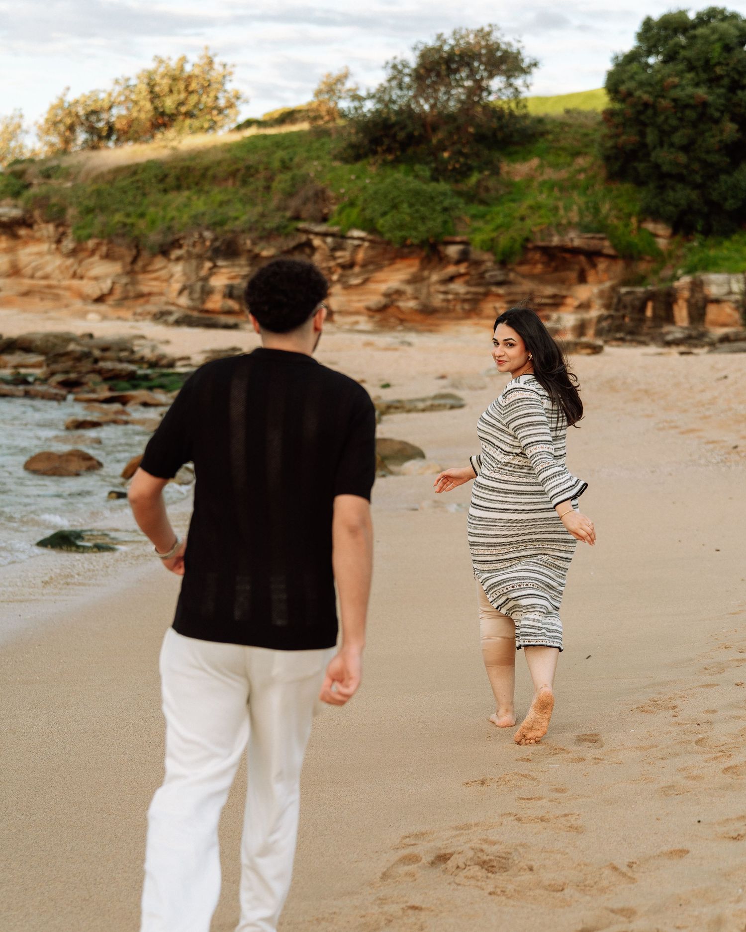 Couple walking on sandy beach at sunset, woman in striped dress looks back smiling at partner.