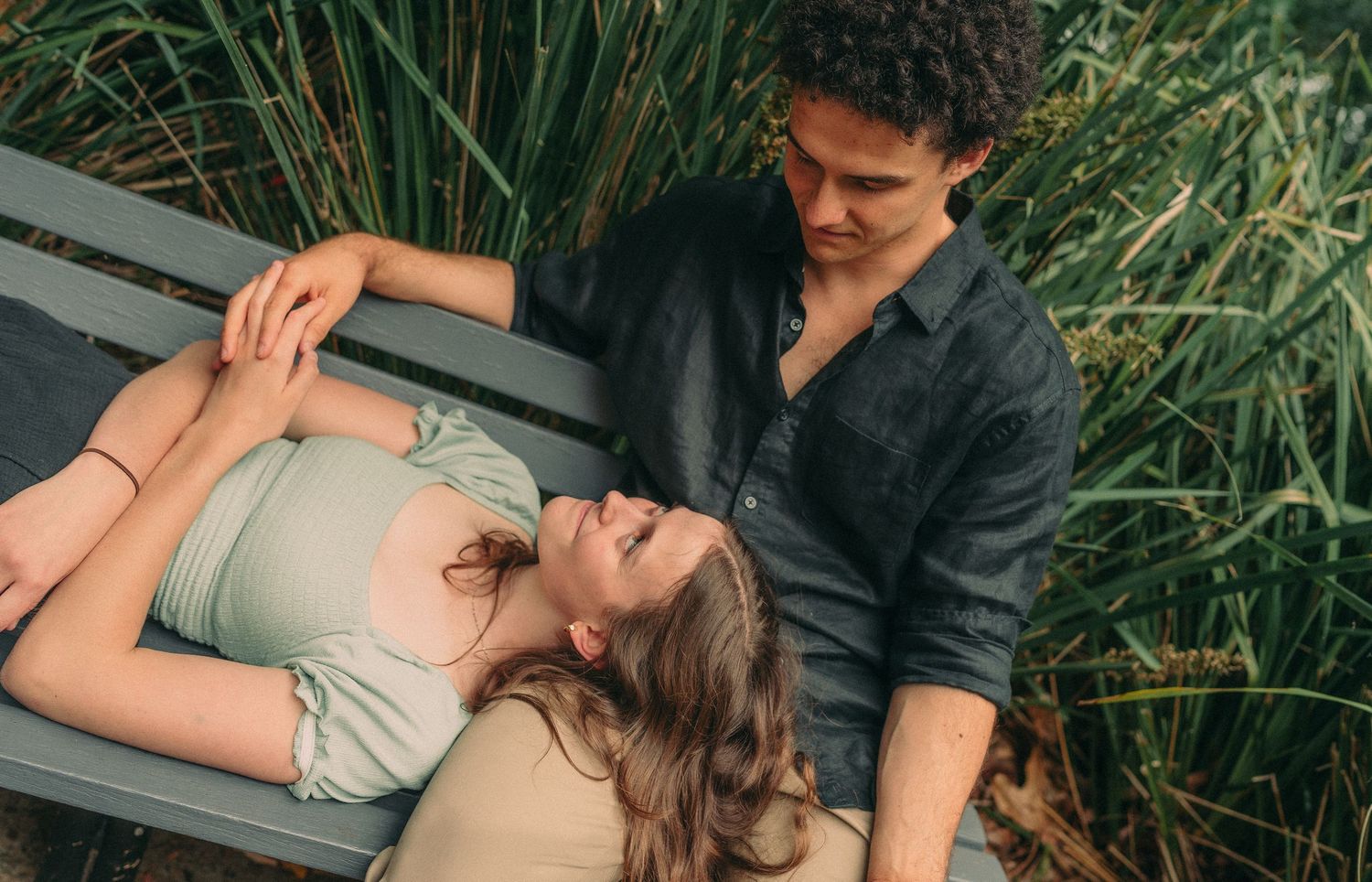 A couple lying on a bench together, smiling and holding hands among tall green grass.