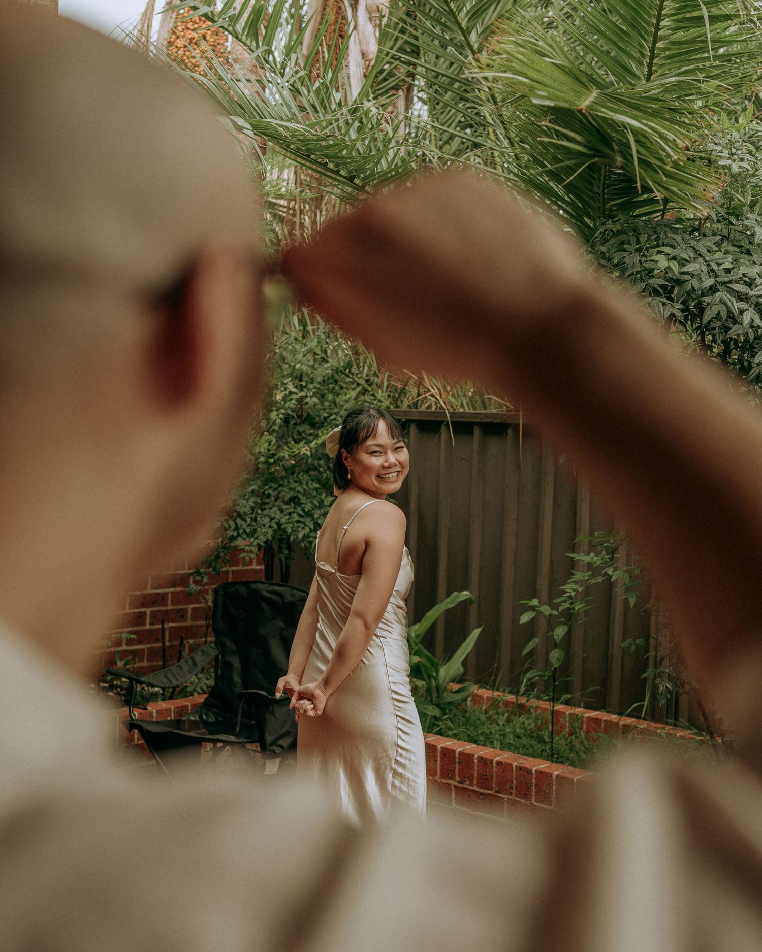 Smiling bride in a satin slip dress holds her partner's hand in a lush green garden.
