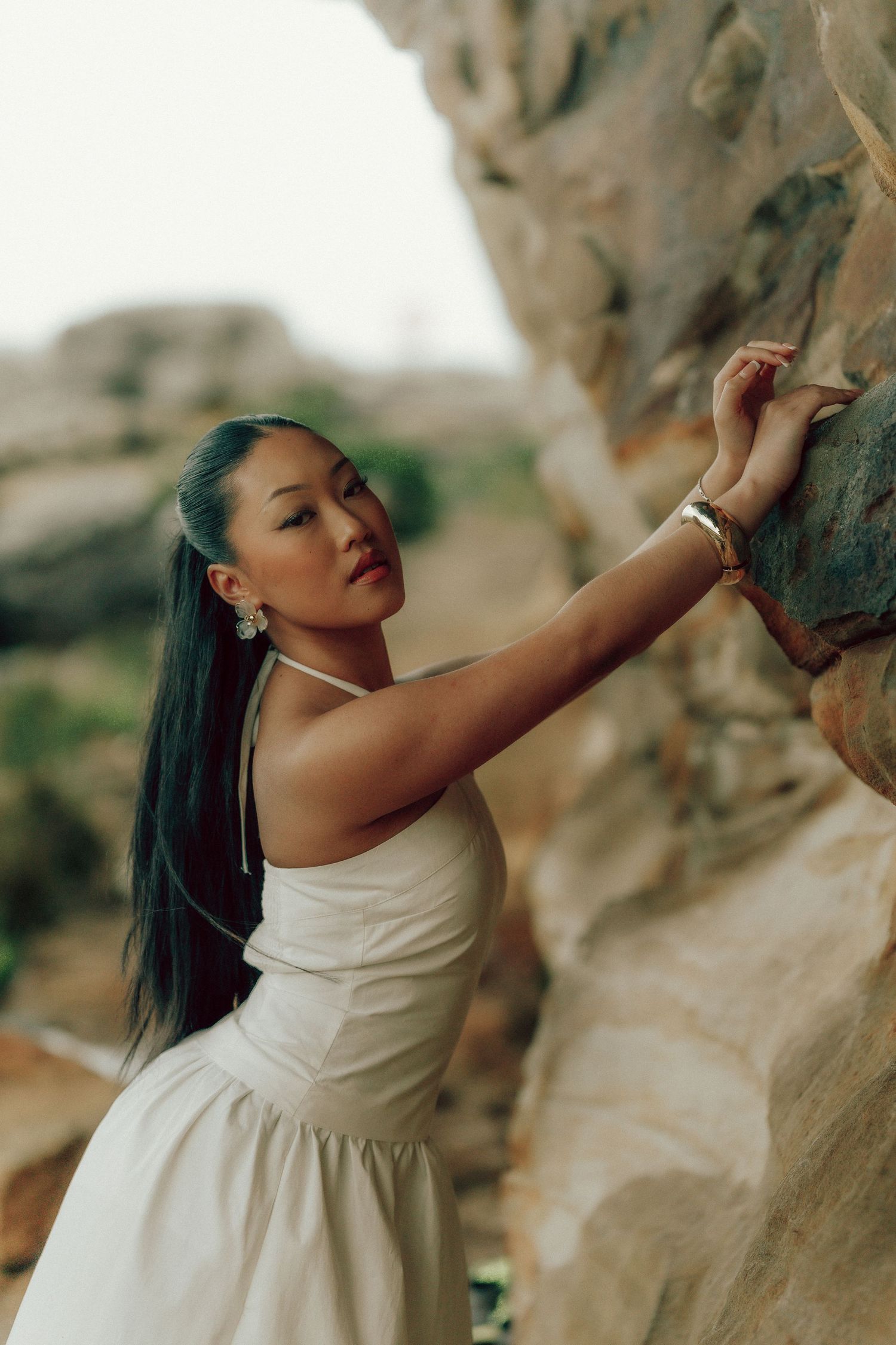 Elegant Asian woman in white halter dress touching rocky cliff face with long dark hair.