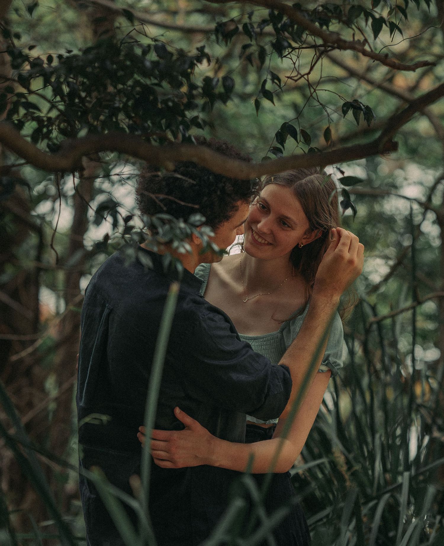 A smiling woman embracing her partner among lush green trees and foliage.