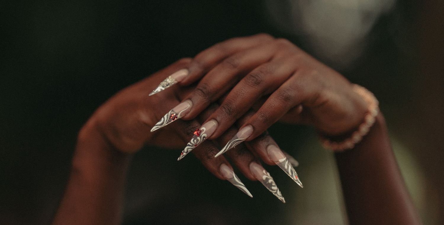 Close-up of dark-skinned hands with long stiletto nails featuring silver glitter nail art and a pearl bracelet.