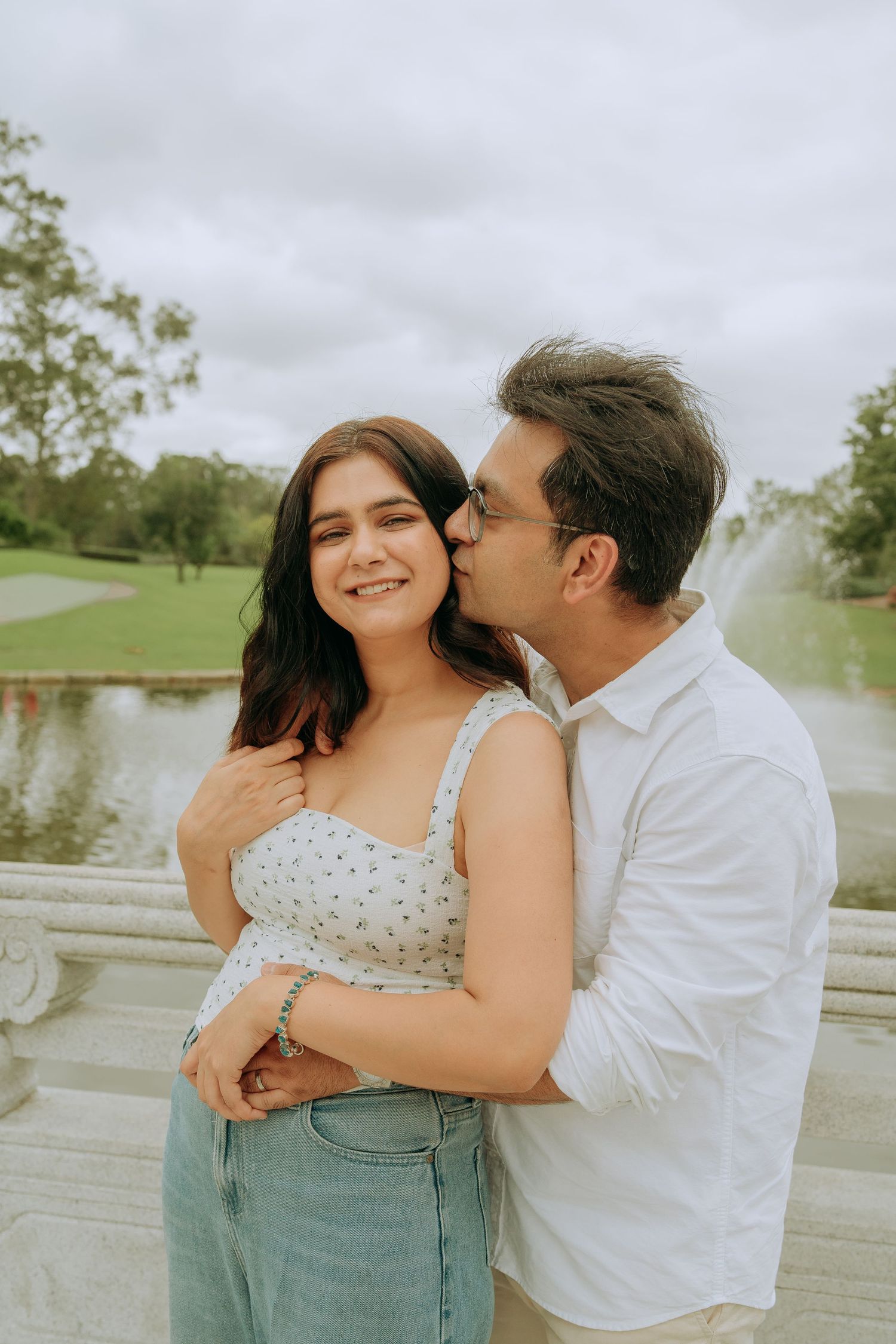 A man tenderly kisses a smiling woman on the cheek while embracing her near a scenic water fountain.