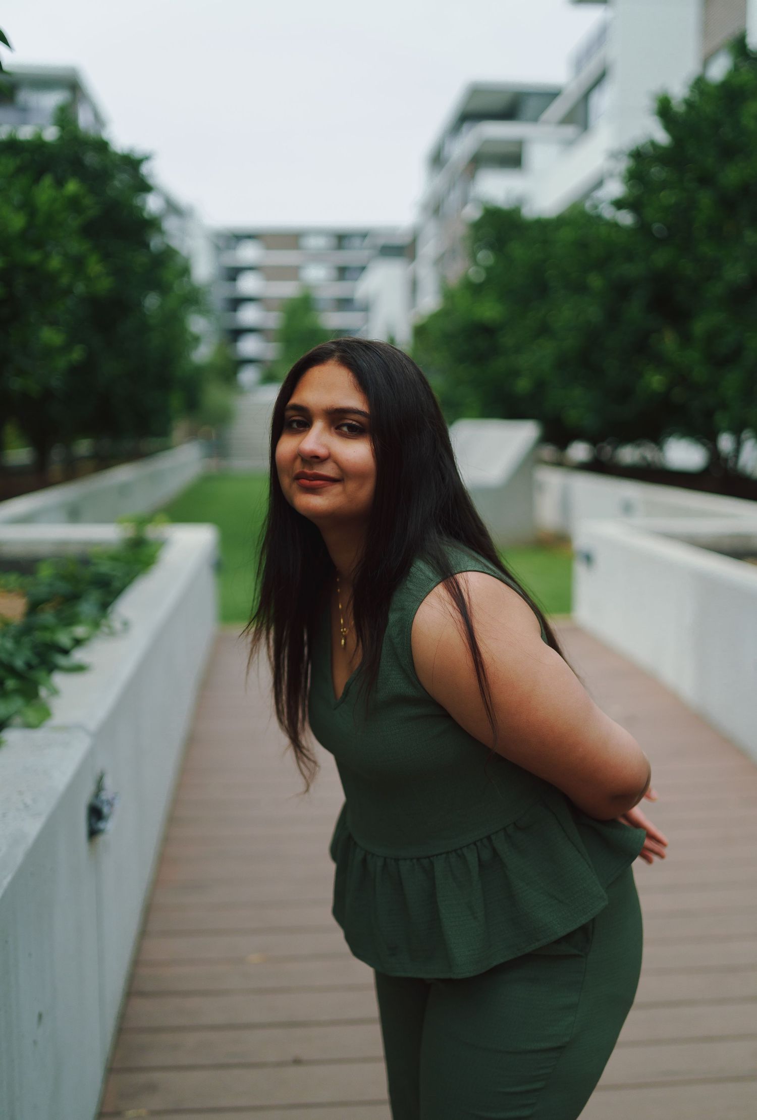 Young woman in green sleeveless outfit poses on wooden walkway surrounded by lush trees and modern buildings.