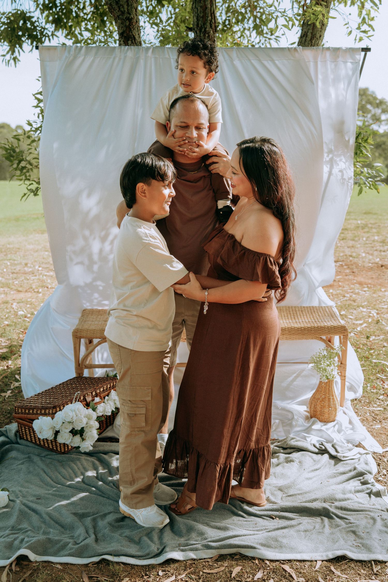 A warm family portrait of parents and two young children posing before a white draped backdrop in a sunny park.